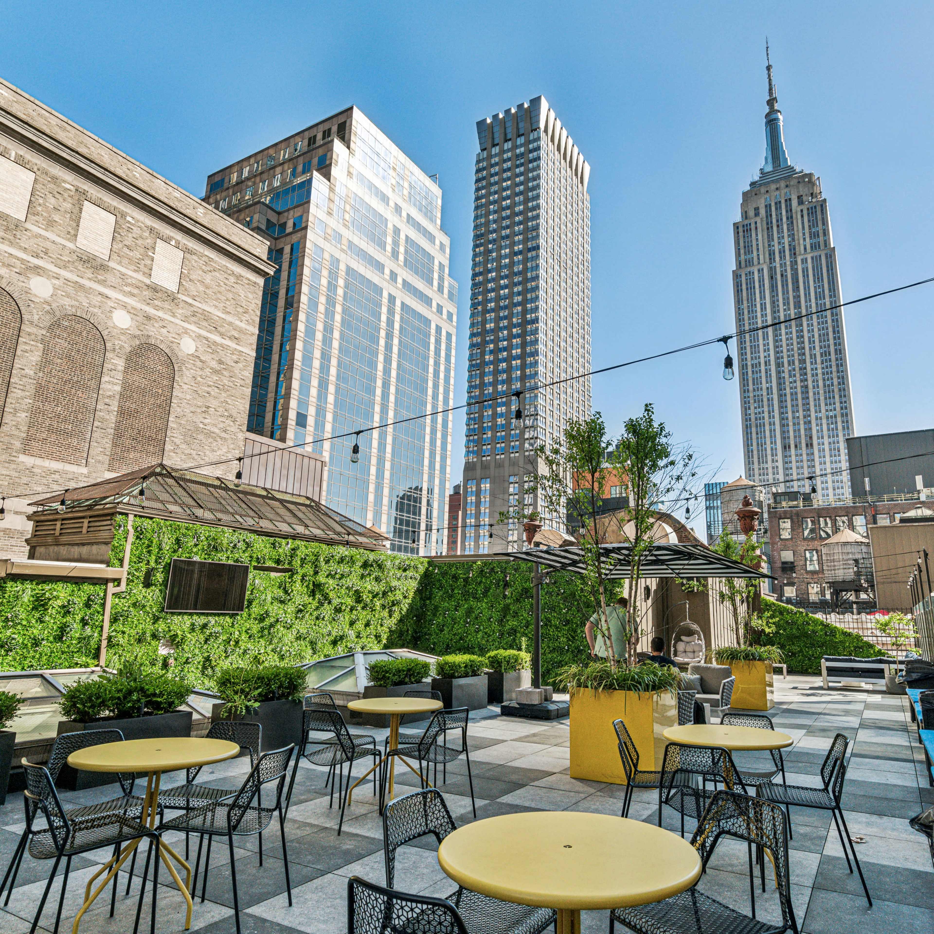 A rooftop terrace with yellow tables and chairs offers a view of the Empire State Building and surrounding skyscrapers.