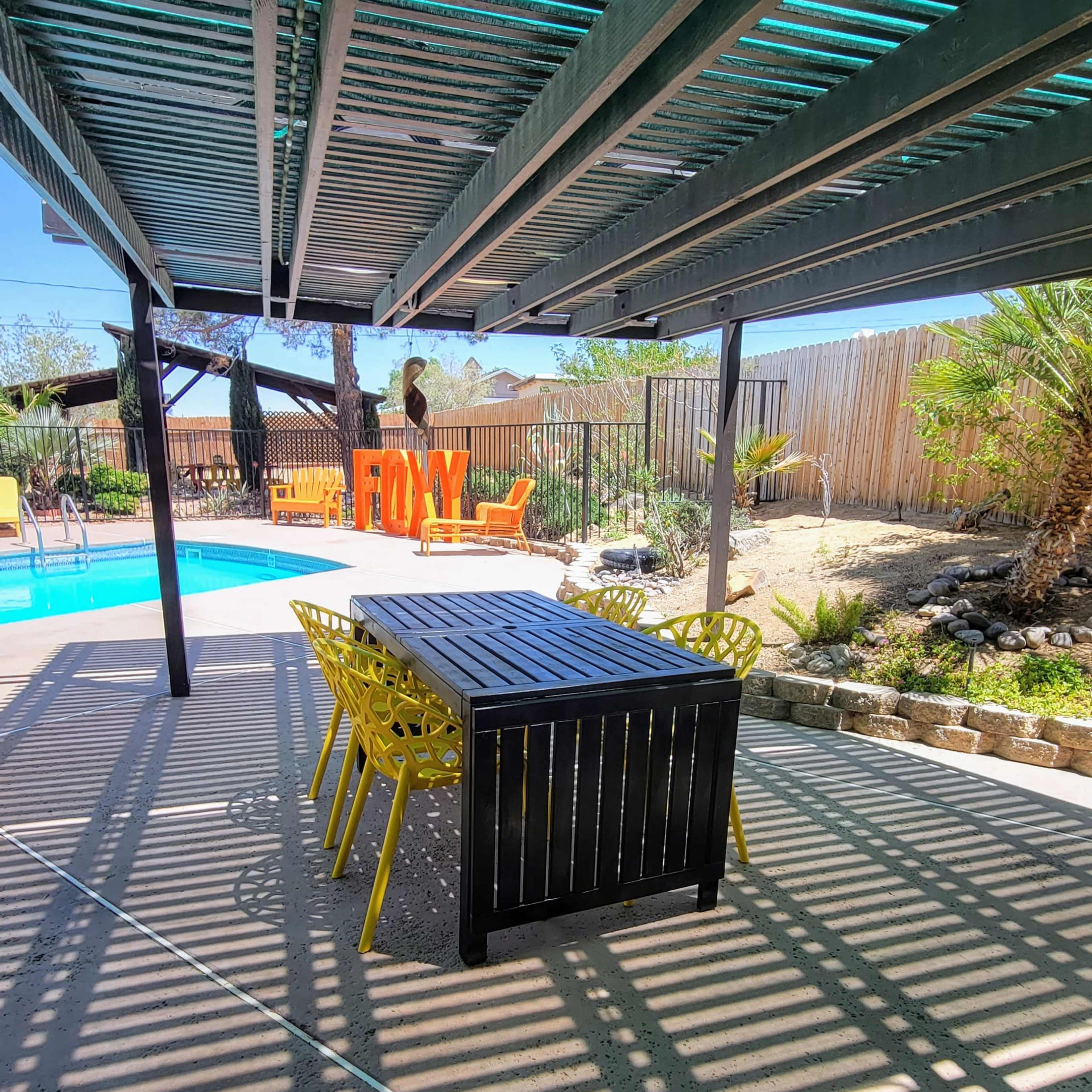 A shaded outdoor dining area features a long black table surrounded by yellow chairs, with a swimming pool and landscaped yard visible in the background.