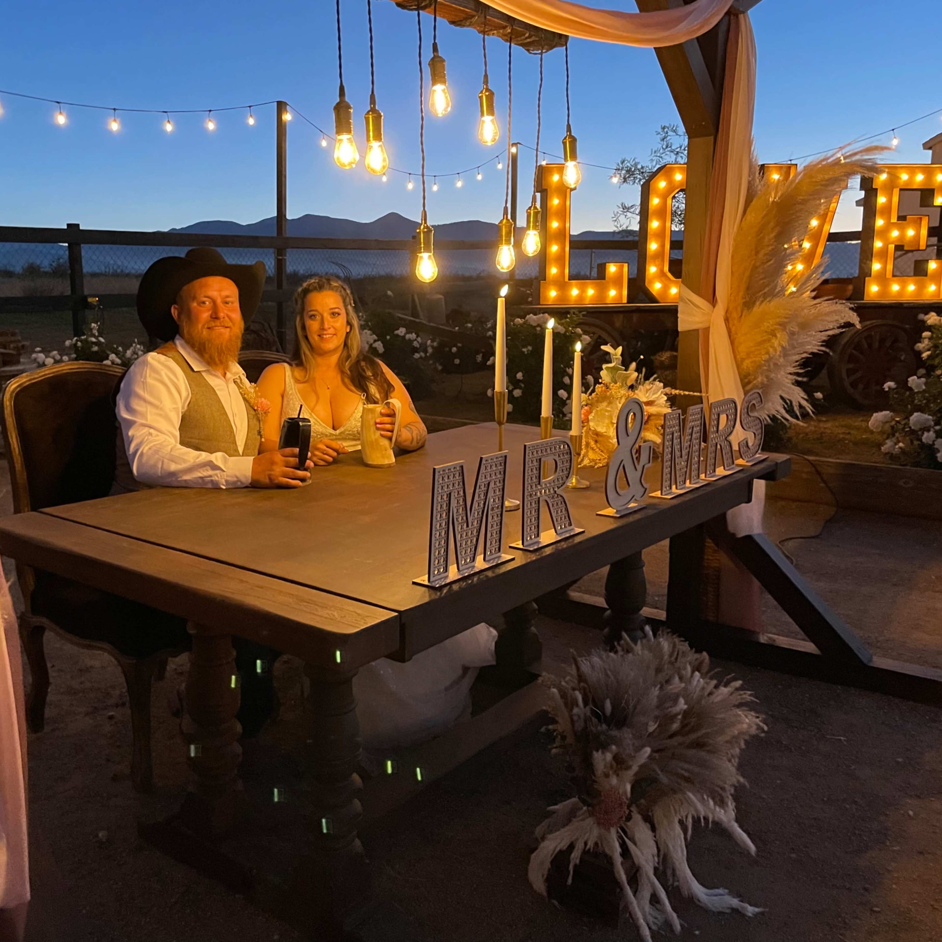 A couple sits at a beautifully decorated table labeled "Mr & Mrs" under string lights at dusk.