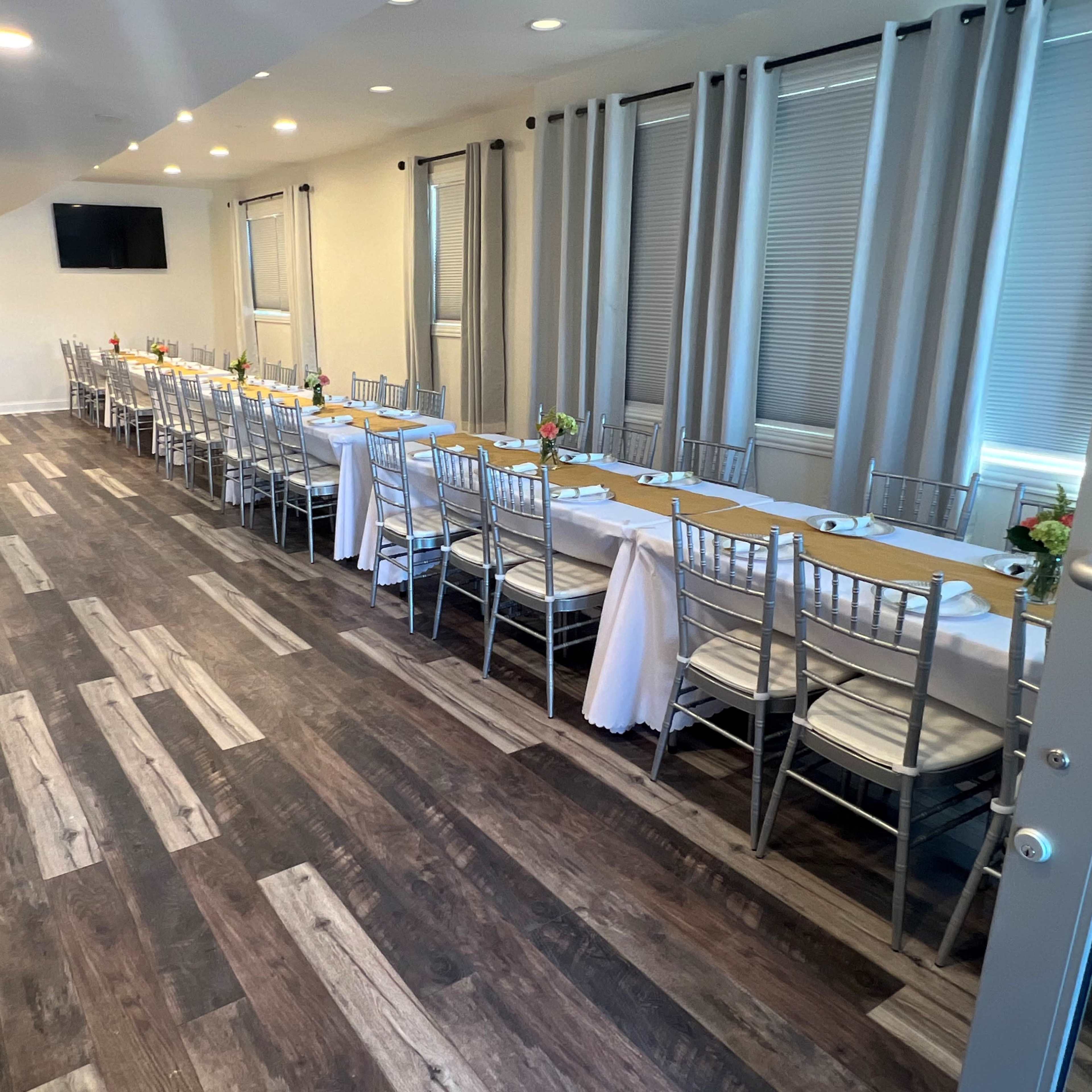 A long dining table is set with white tablecloths and chairs in an elegantly arranged hallway with light-colored walls and wood flooring.