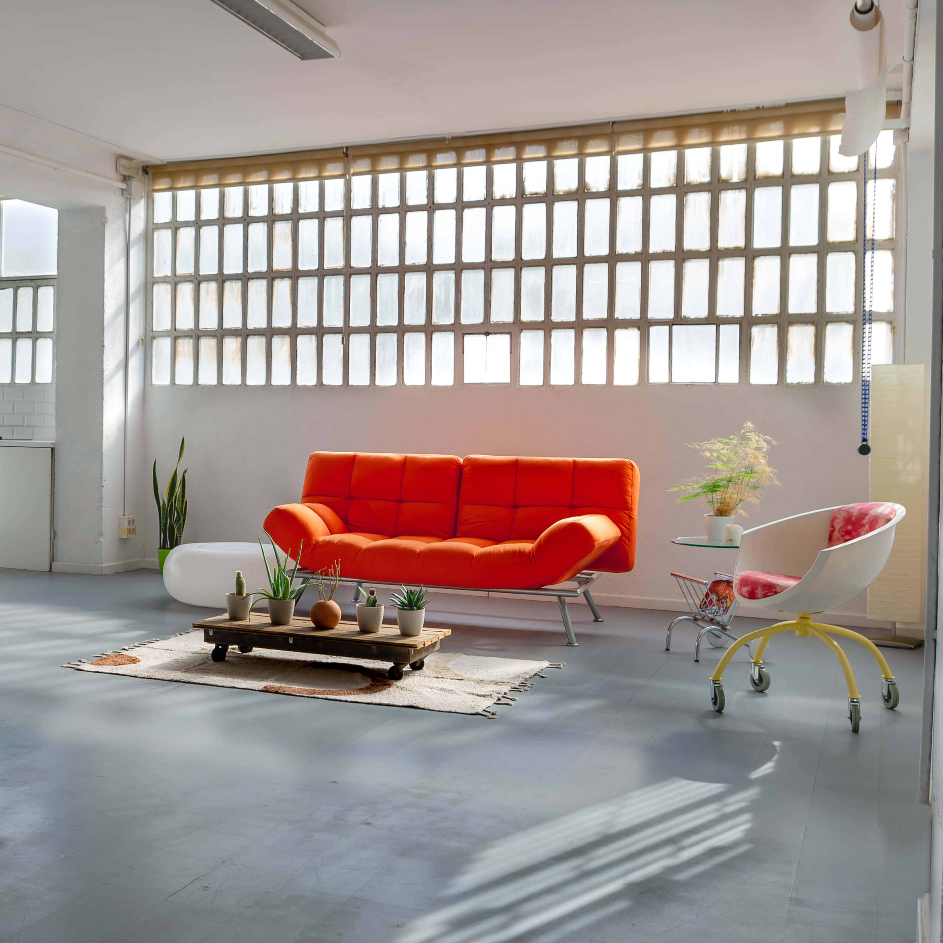 A bright, open living space features an orange couch, a wooden coffee table with decorative items, and a small kitchen area in the background.