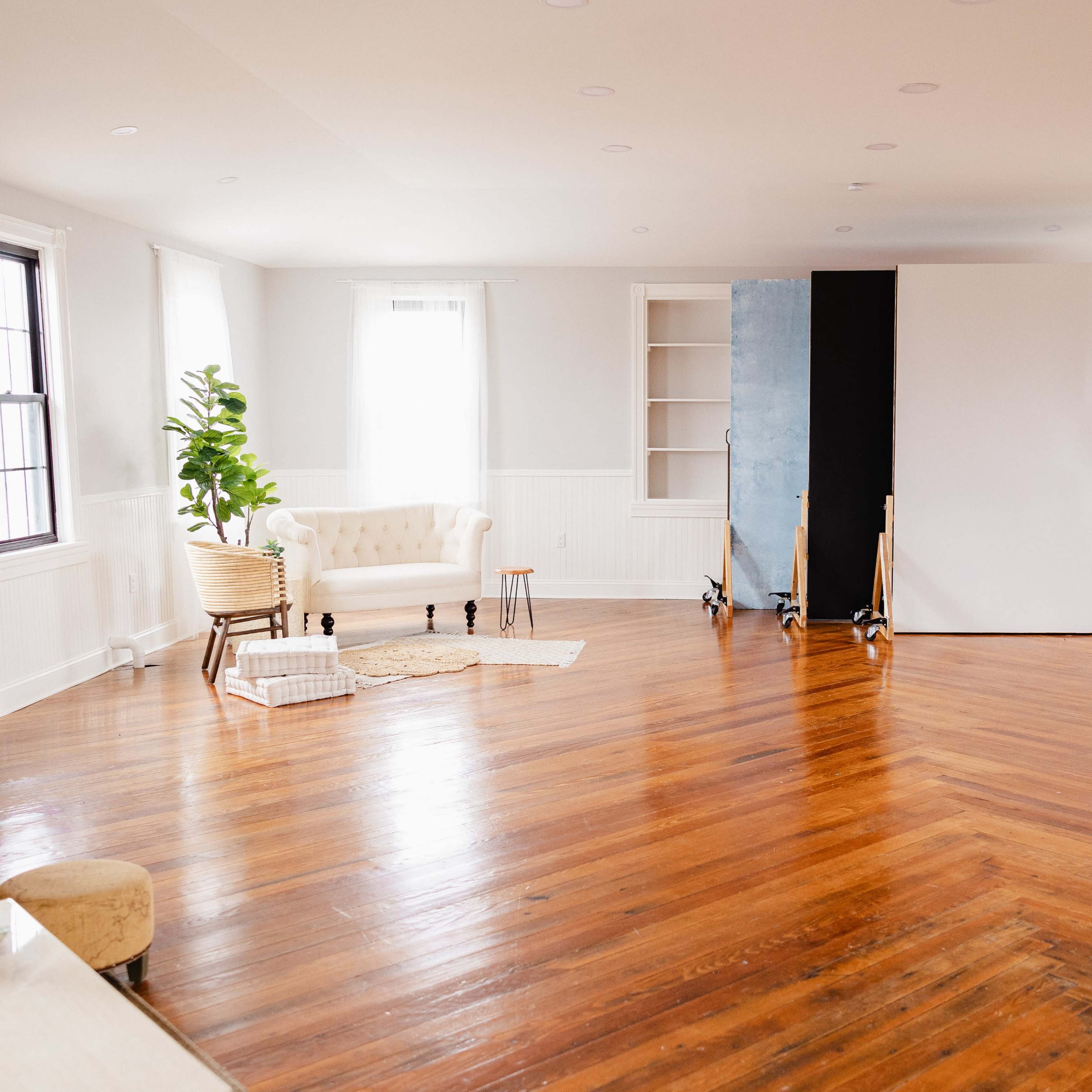 A spacious, well-lit room with wooden flooring and a white couch, featuring a backdrop setup against a wall.
