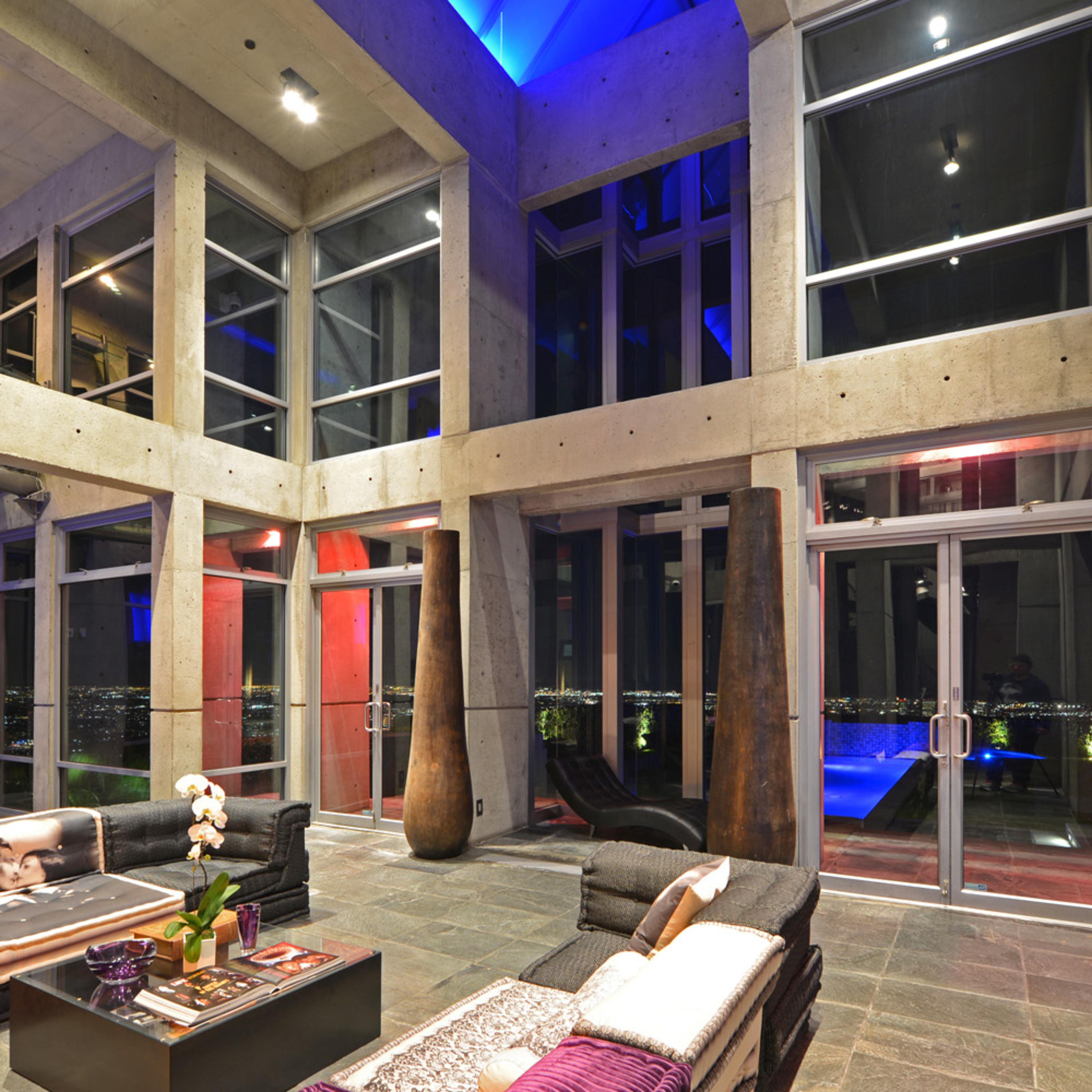 The interior of a modern, open-concept living space features large glass windows, concrete walls, and minimalistic furniture arranged around a stone floor, with views of city lights beyond.