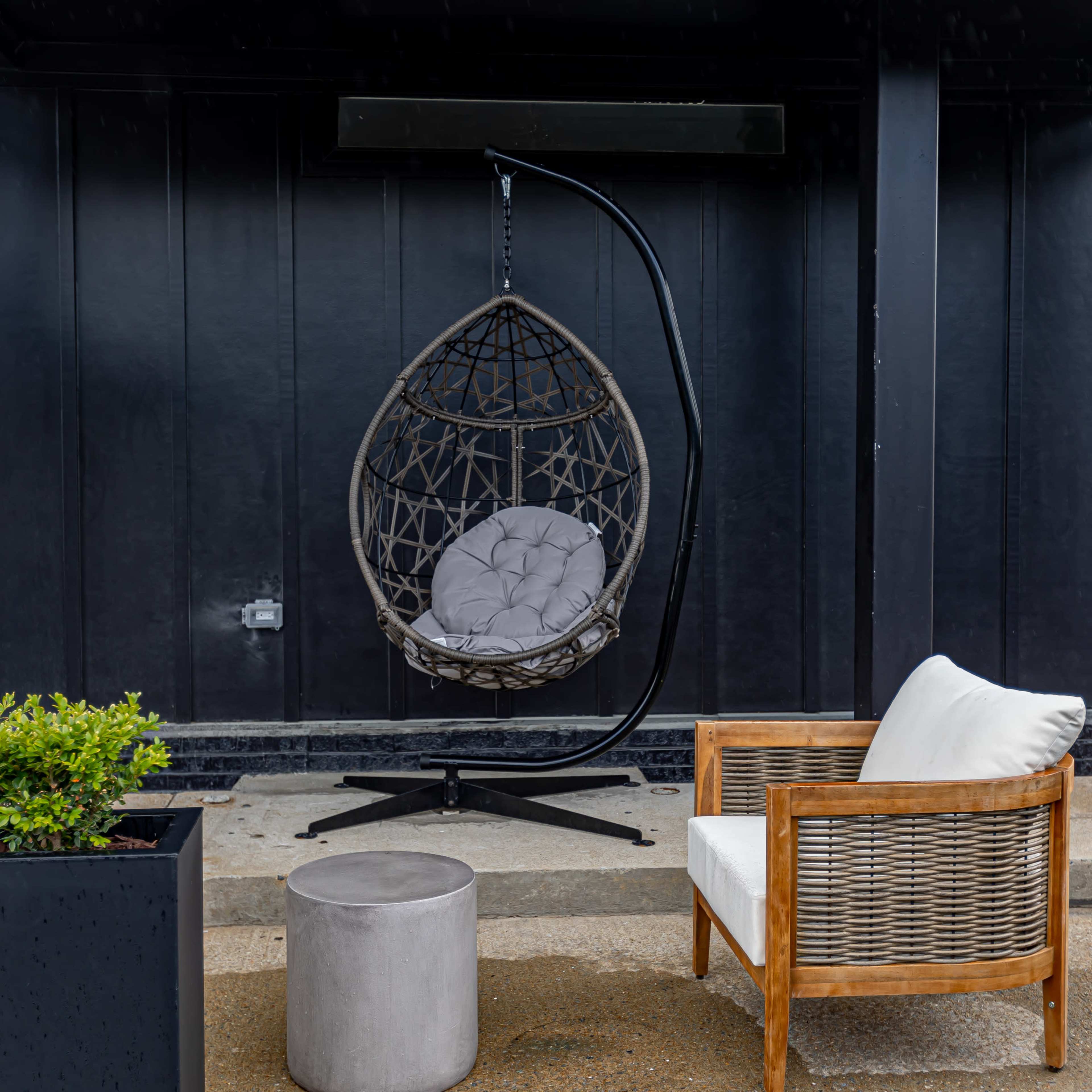 A hanging wicker chair is suspended from a black structure beside a small table and a wooden armchair in a covered outdoor area.
