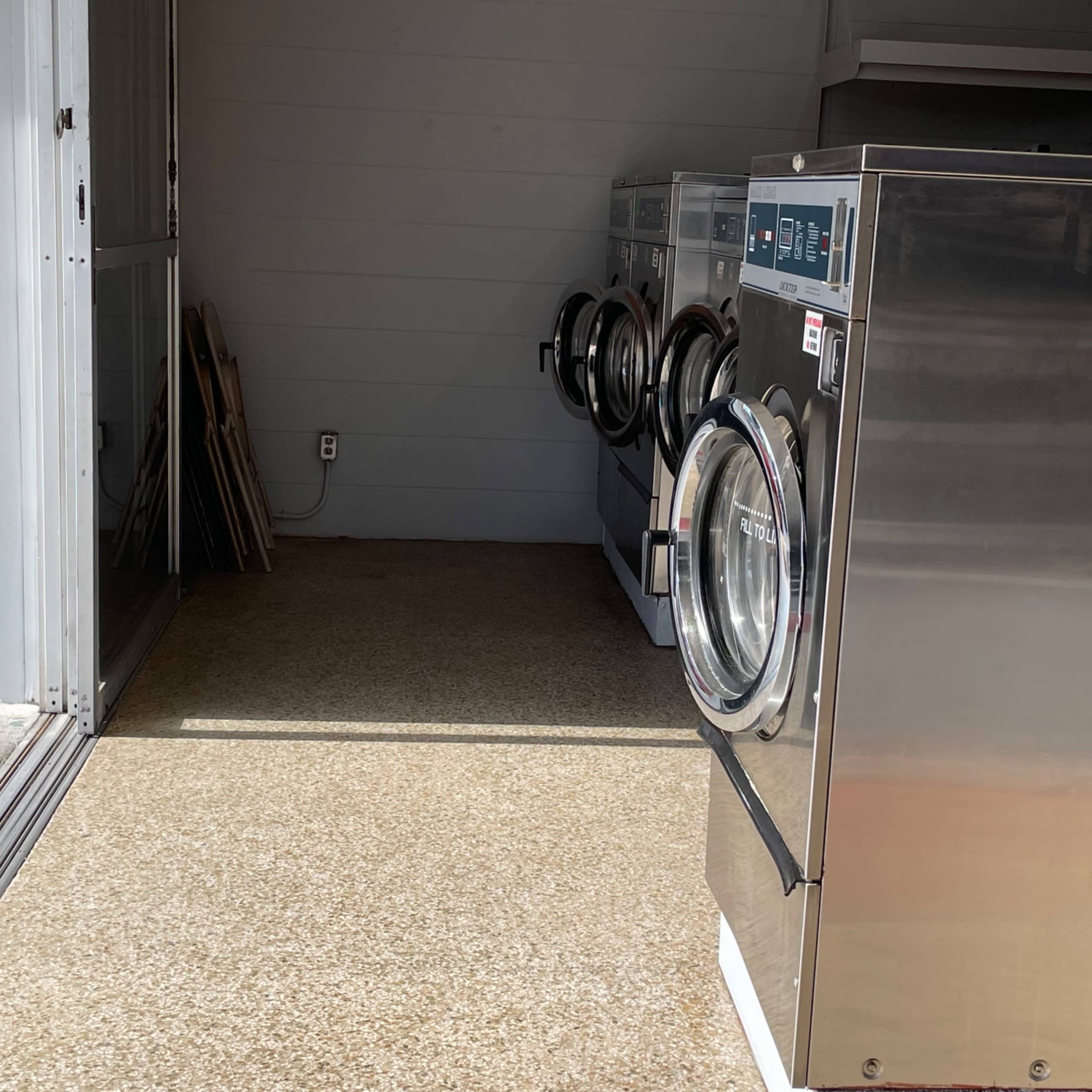 A row of commercial washing machines placed in an indoor laundry area with a concrete floor and a wooden bench in the background.