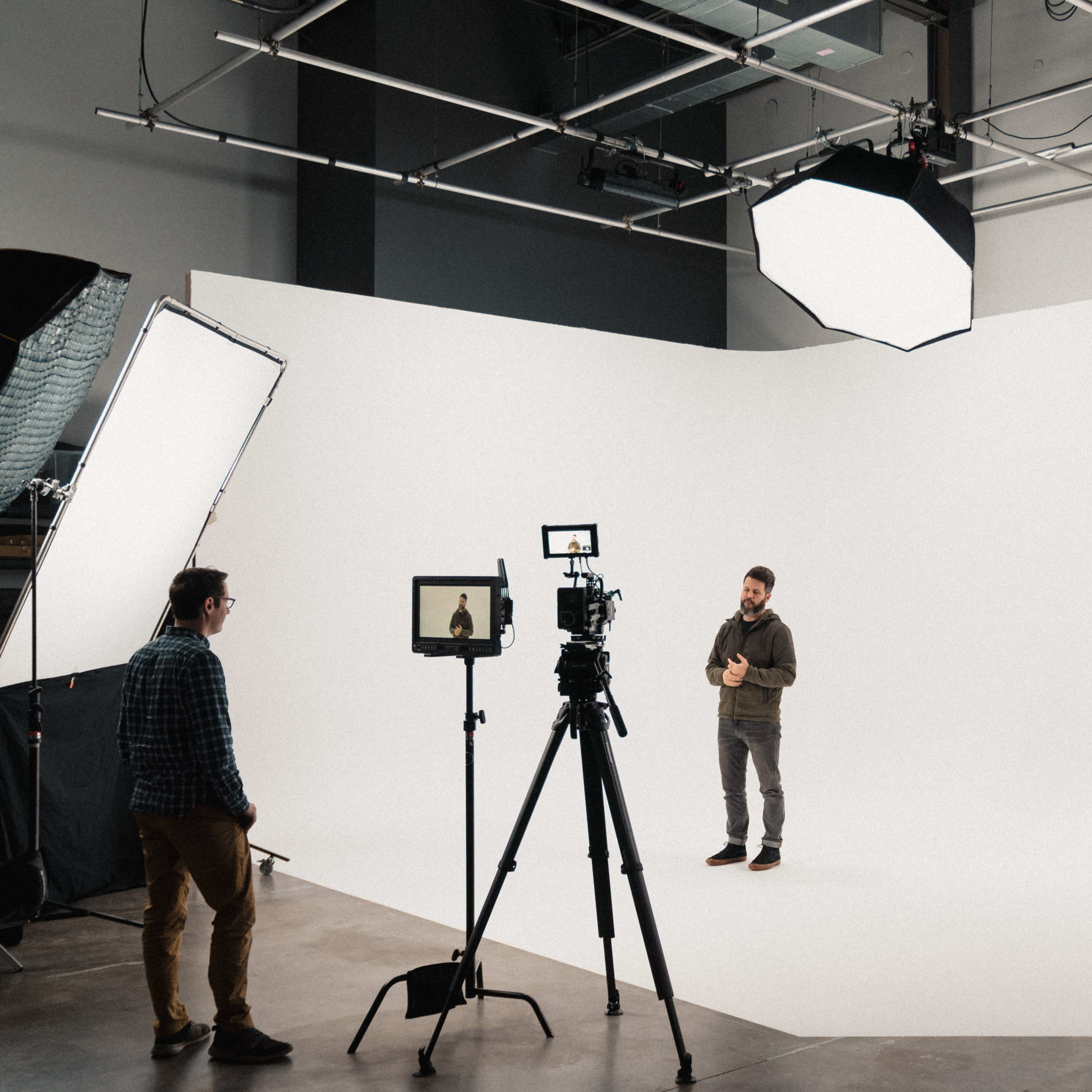 A man stands on a white backdrop speaking into a camera, while another person monitors the setup with a tablet.