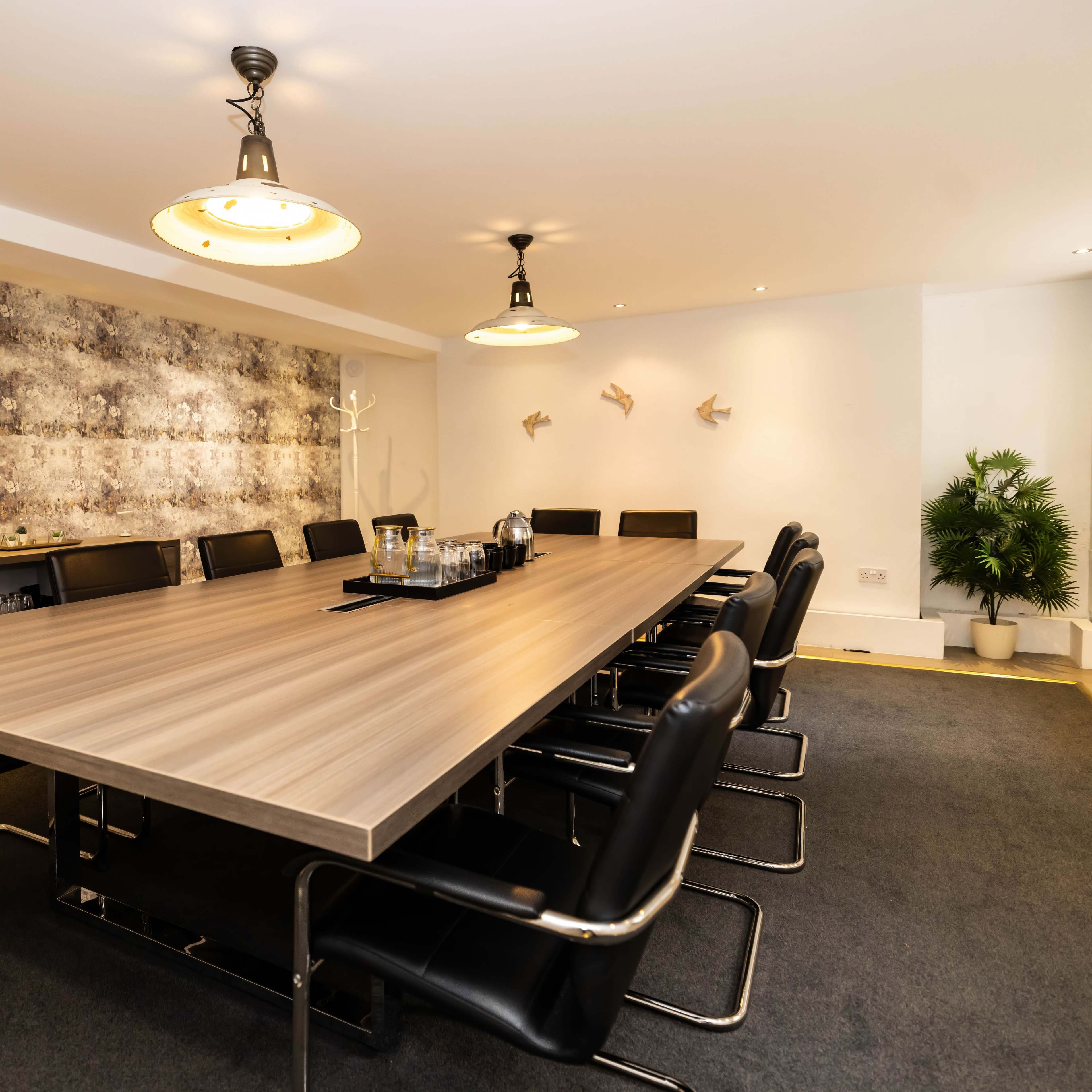 A long wooden conference table with black chairs is set in a well-lit meeting room featuring gray textured wallpaper and a potted plant.