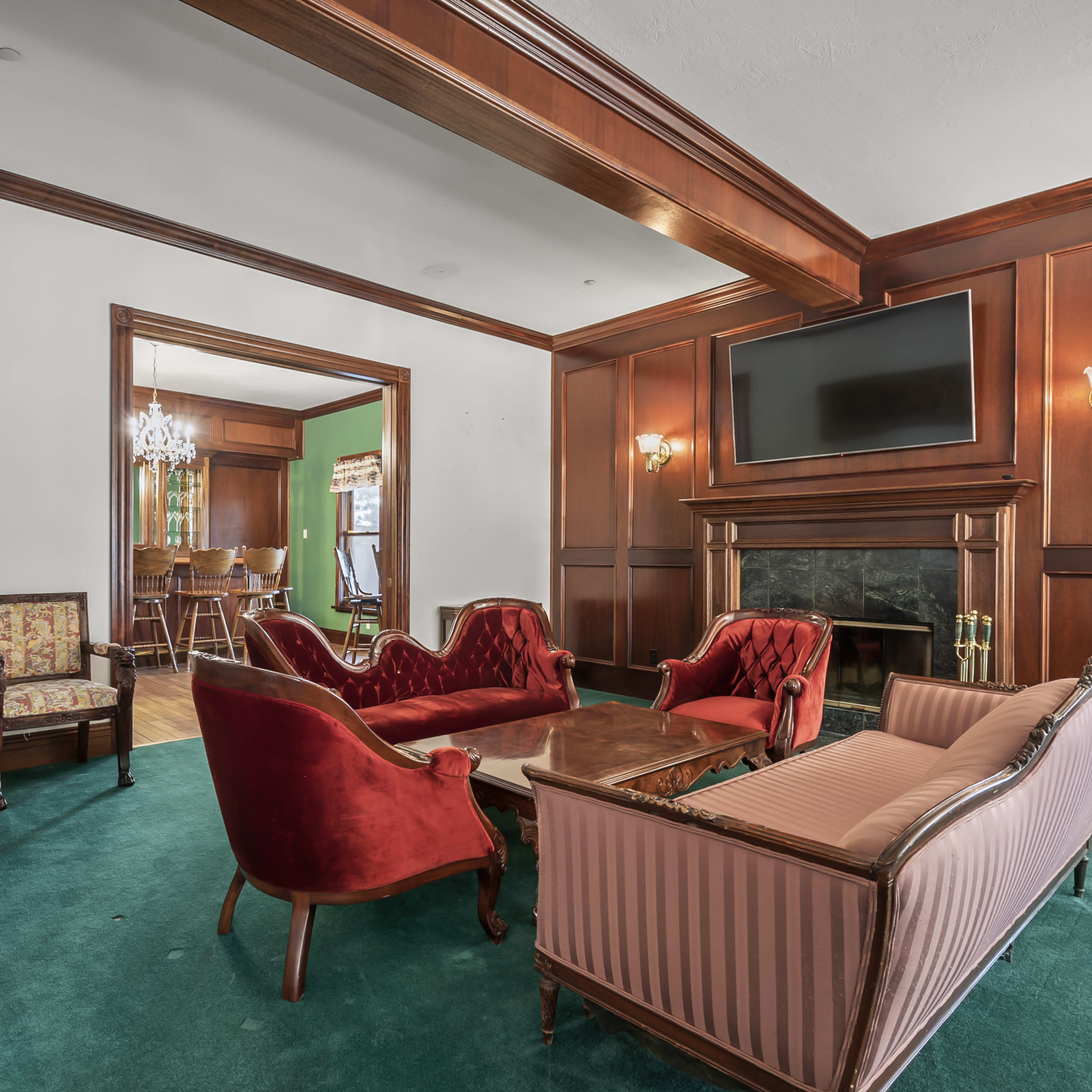 The room features a green carpet, wooden paneling, a large television mounted above a fireplace, and seating arranged with two red sofas and a pair of patterned armchairs.