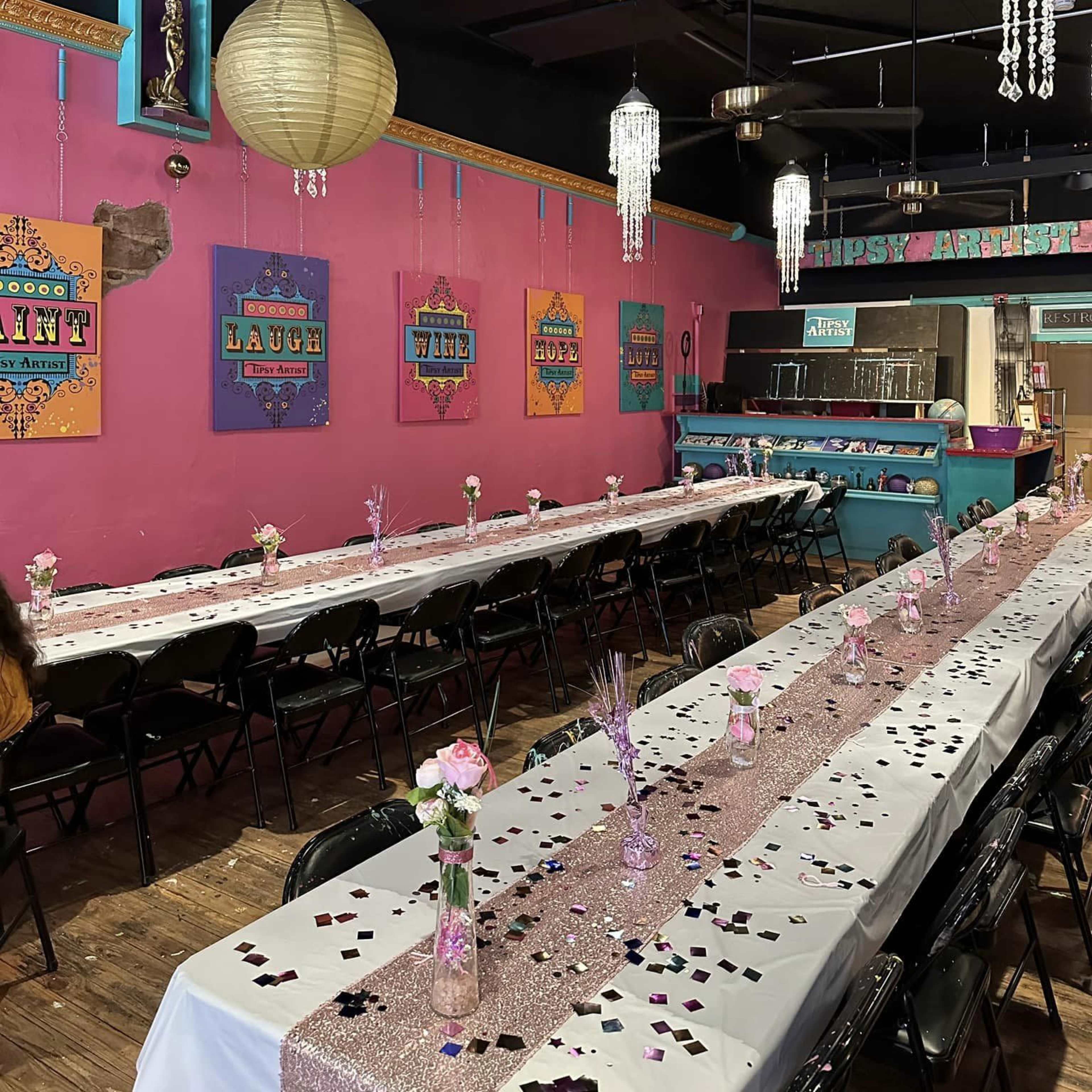 A colorful dining area is set up with long tables adorned with white tablecloths, centerpieces of flowers, and scattered confetti, surrounded by decorative lanterns and vibrant wall art.