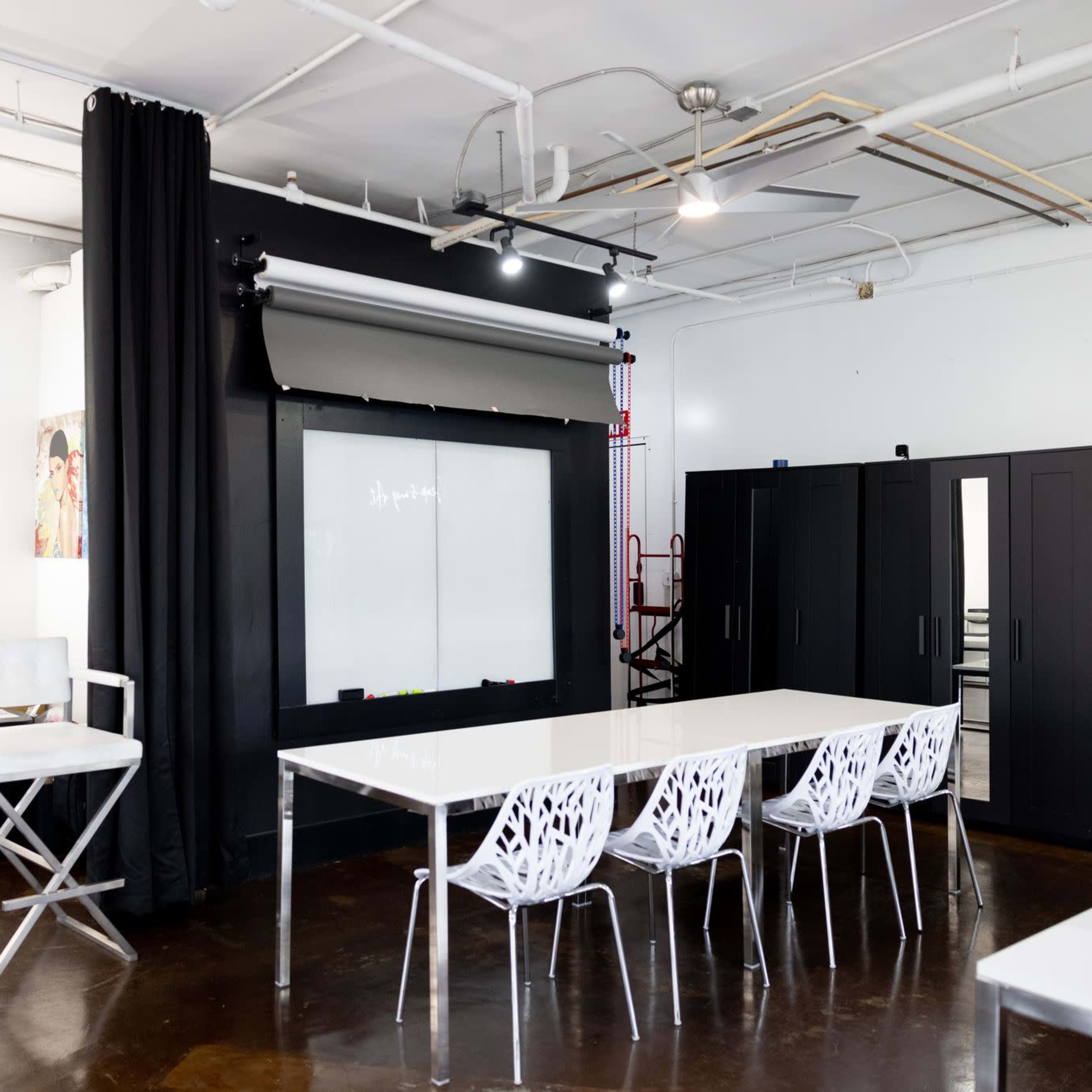 A modern studio space with a large black backdrop, a whiteboard, and a long white table surrounded by decorative chairs.