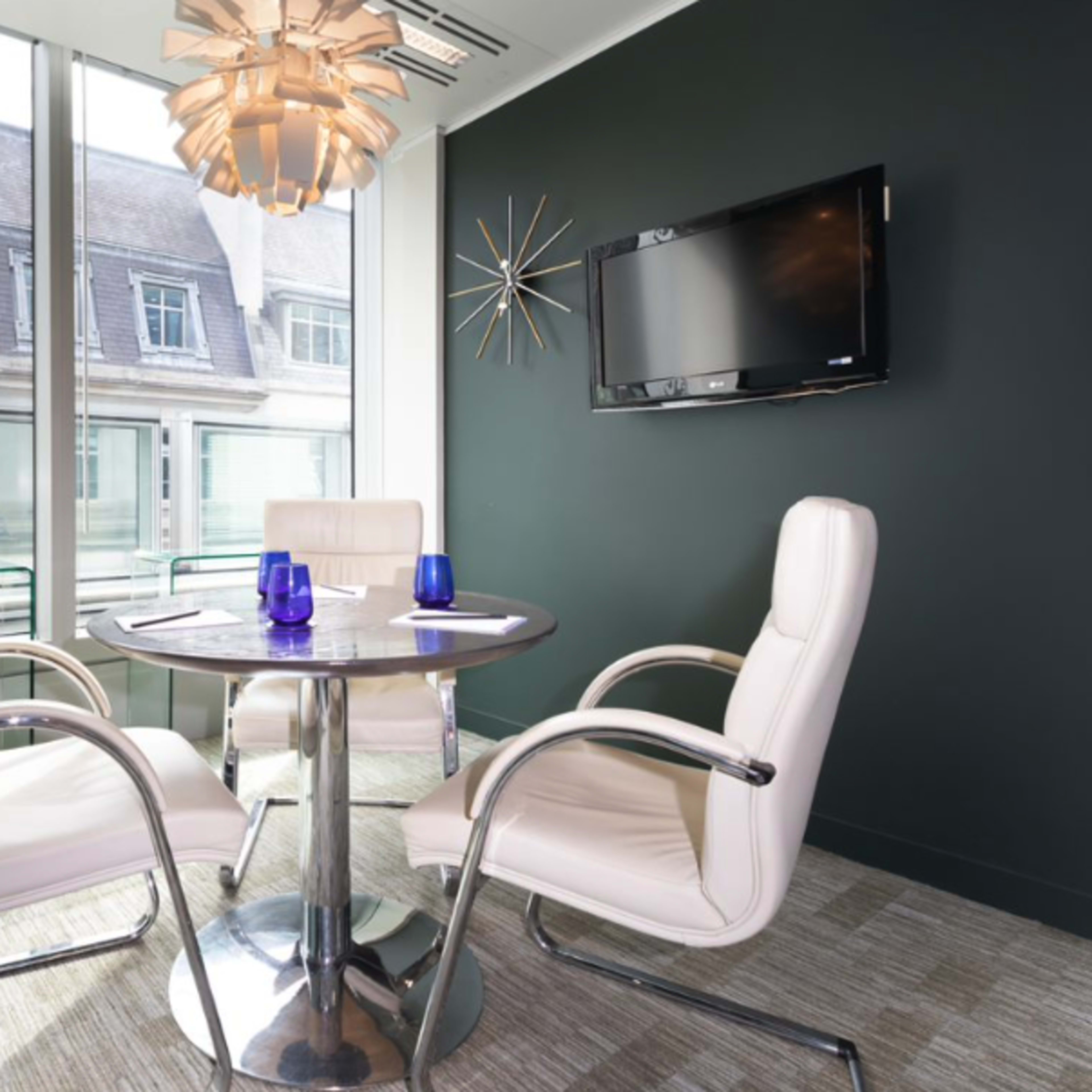 A modern meeting room features a round table with four white chairs, large windows overlooking a building exterior, and decorative elements including a pendant light and a wall-mounted TV.