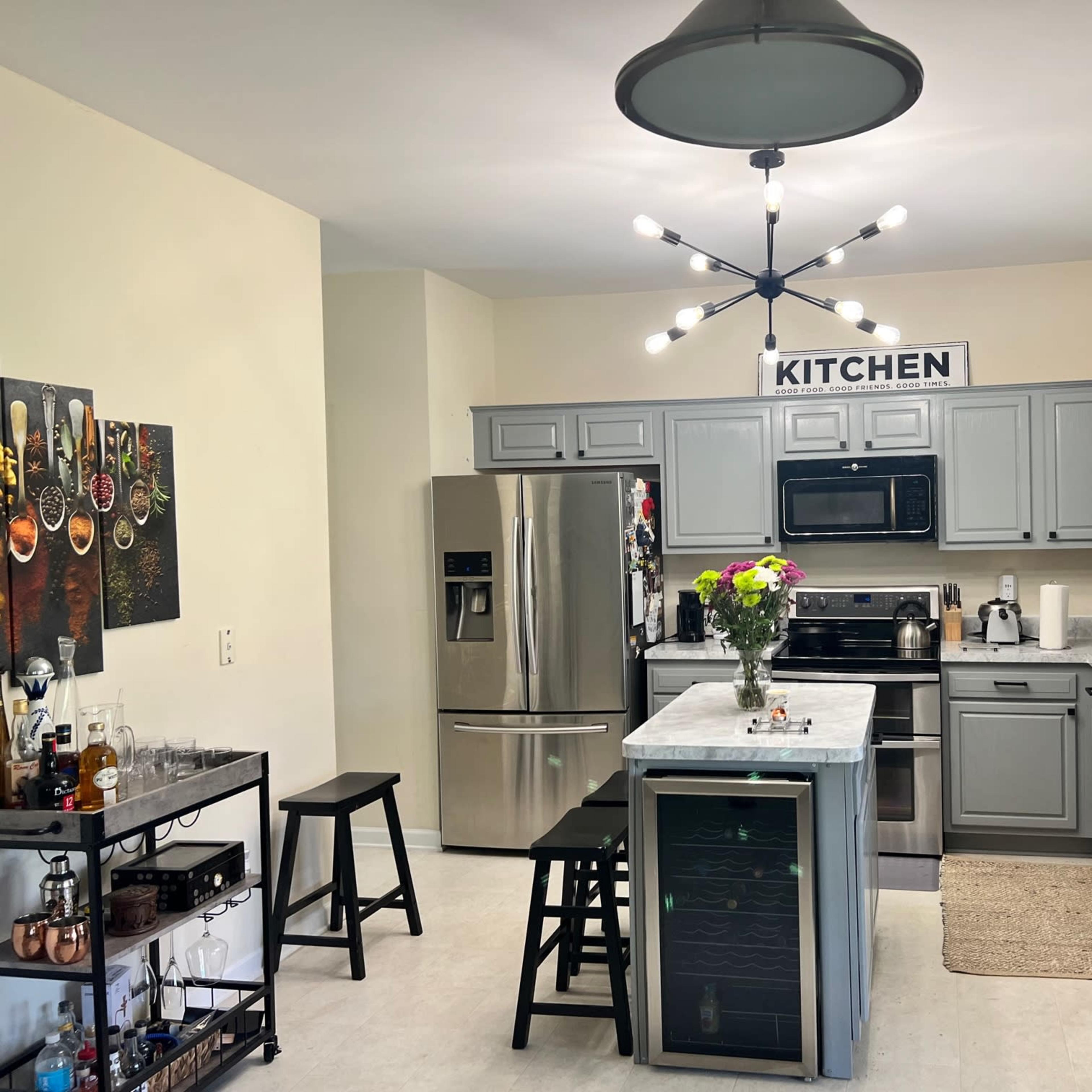 A modern kitchen features gray cabinets, stainless steel appliances, and a central island with seating, illuminated by a decorative light fixture.