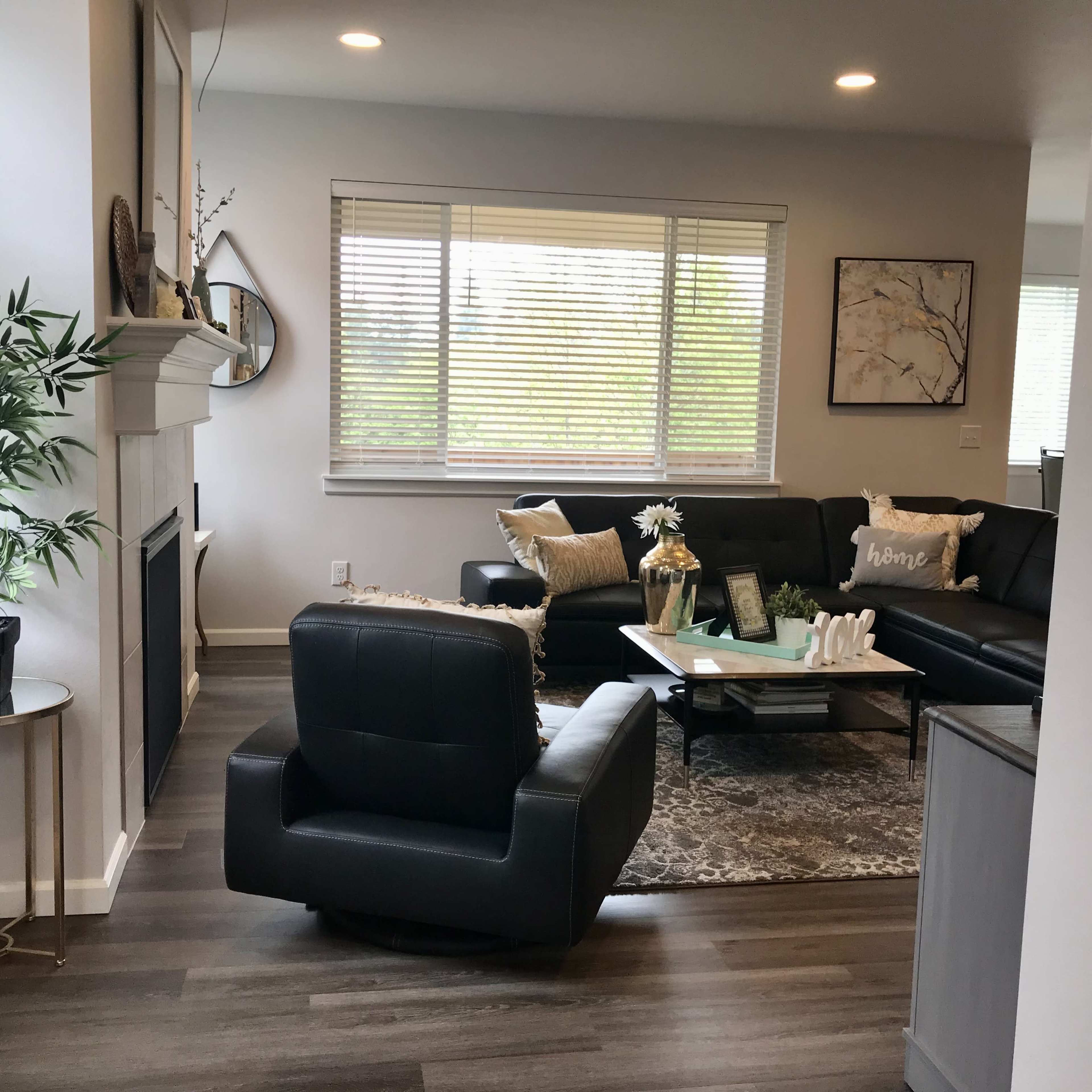 A modern living room features a black leather sofa, a swivel chair, a coffee table with decorative items, and large windows allowing natural light.