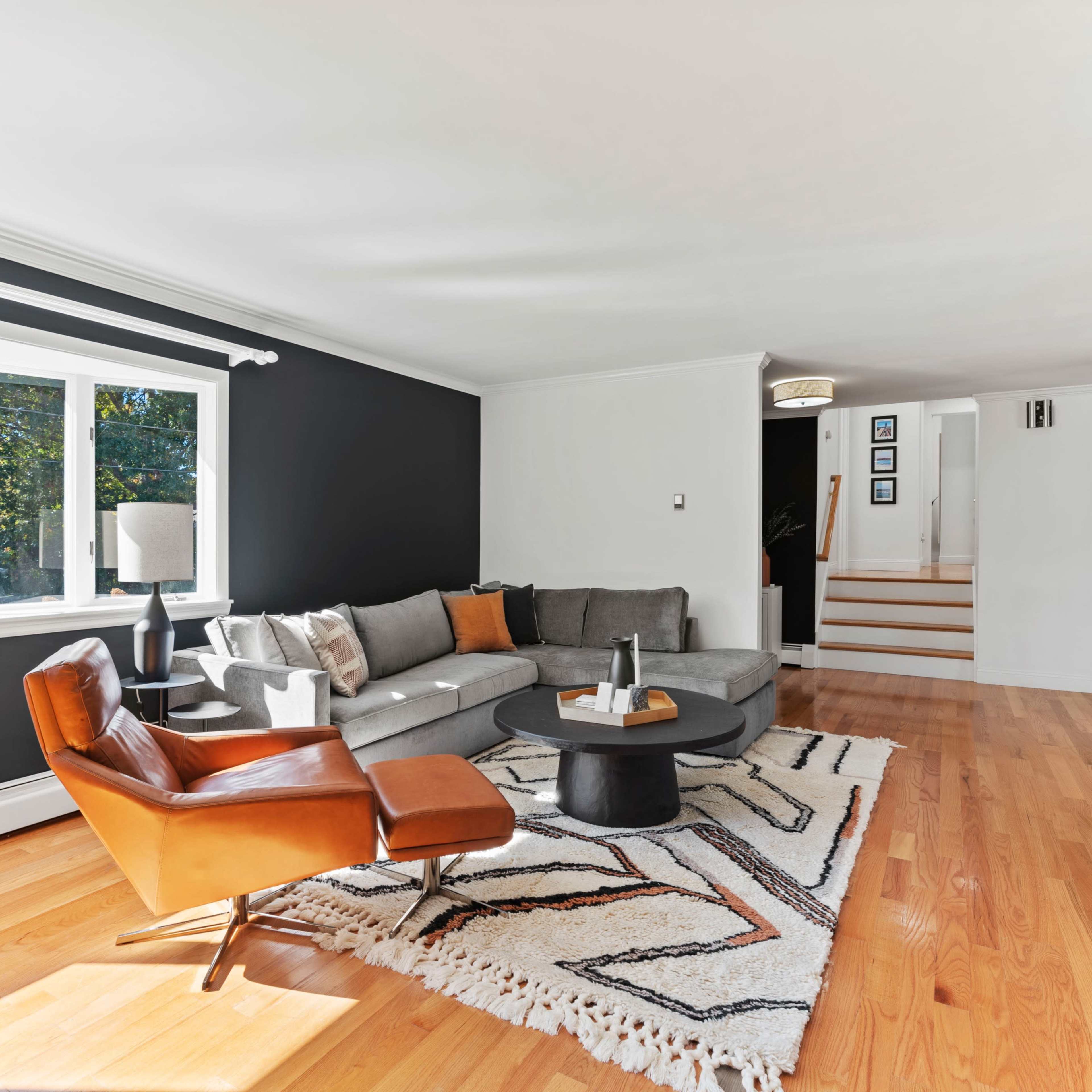 A living room features a gray sectional sofa, an orange accent chair, a circular coffee table, and large windows allowing natural light to enter.