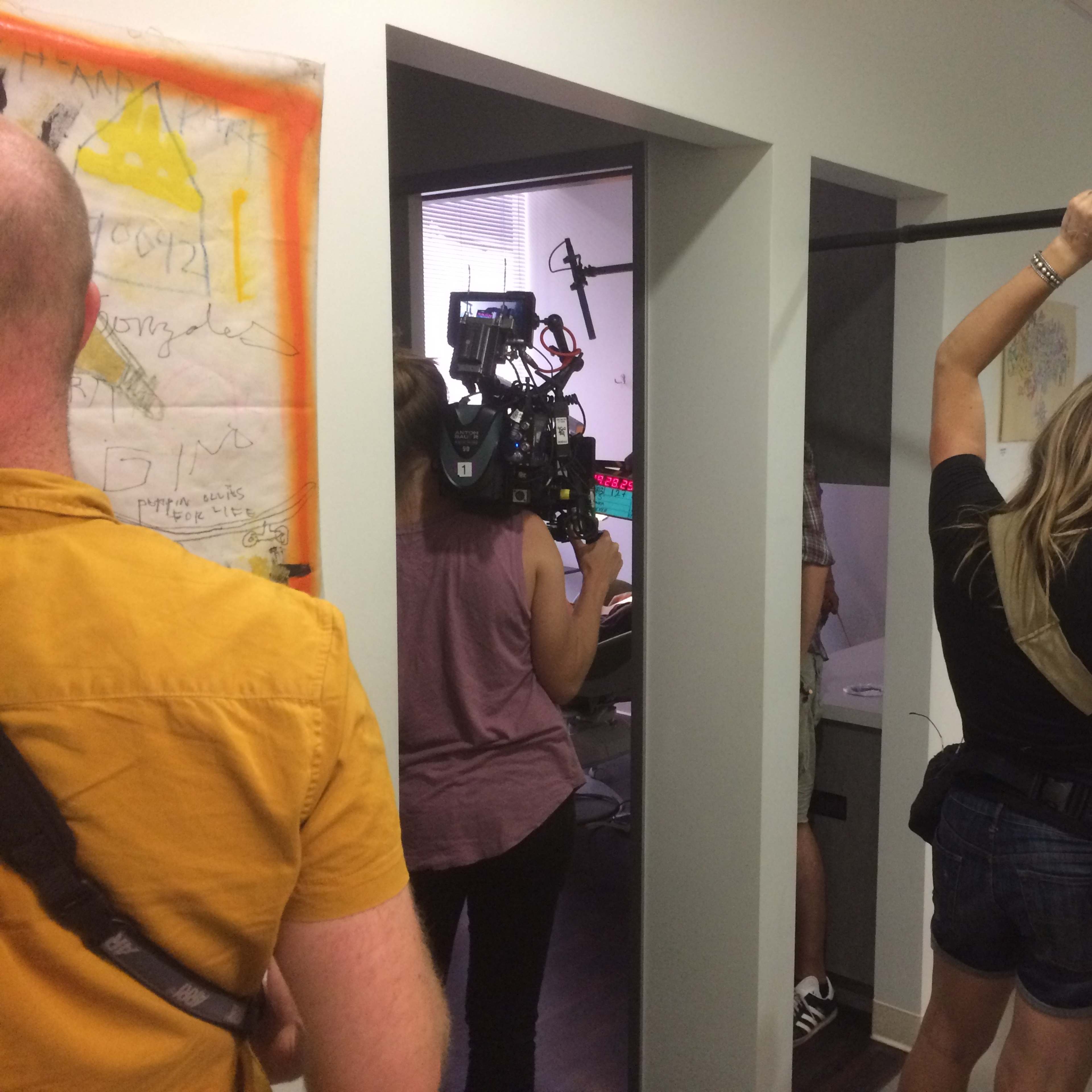 A film crew is set up in a hallway, with one person holding a boom microphone while others operate a camera and adjust equipment.