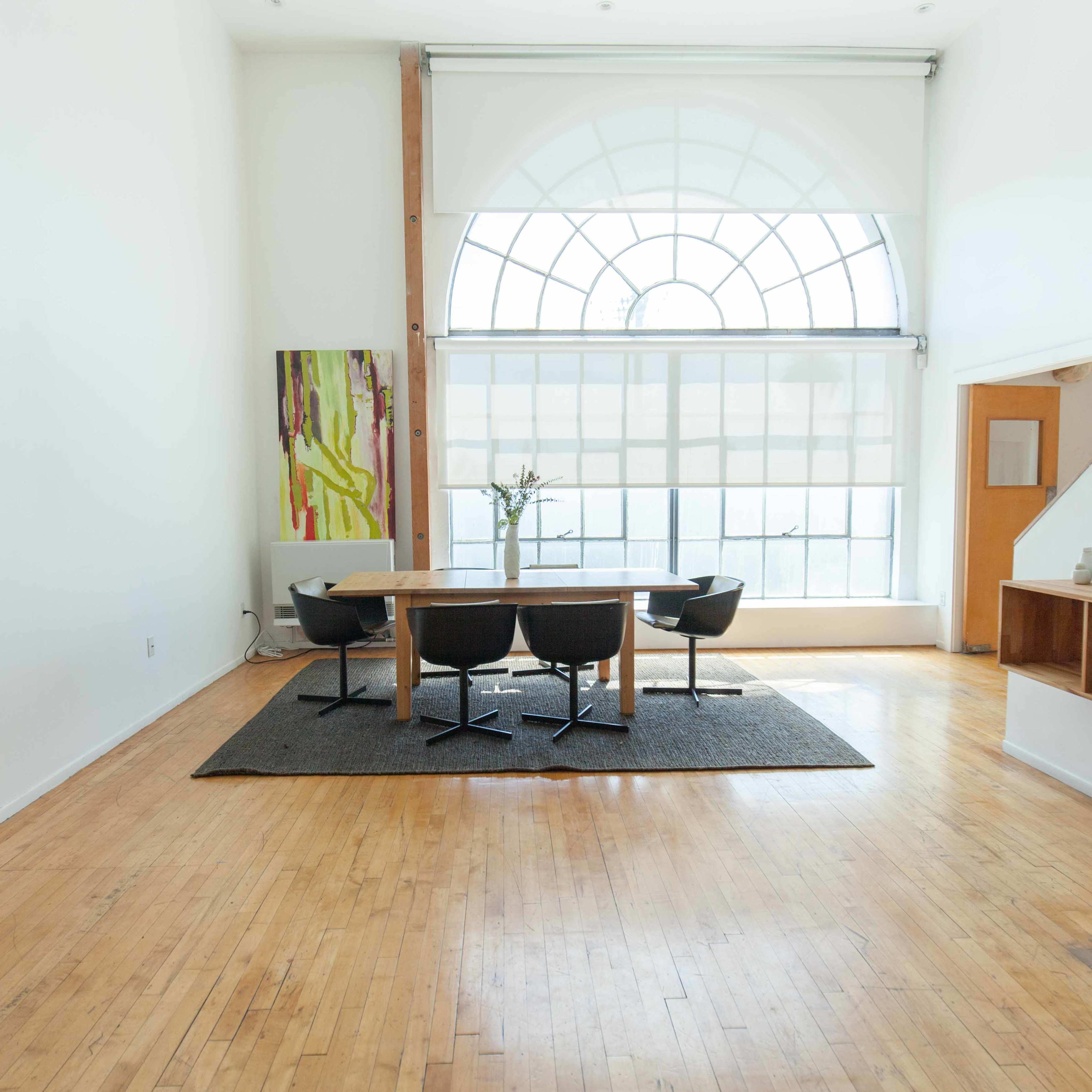 A bright, minimalist interior featuring a wooden table surrounded by black chairs on a rug, with a large arched window in the background.