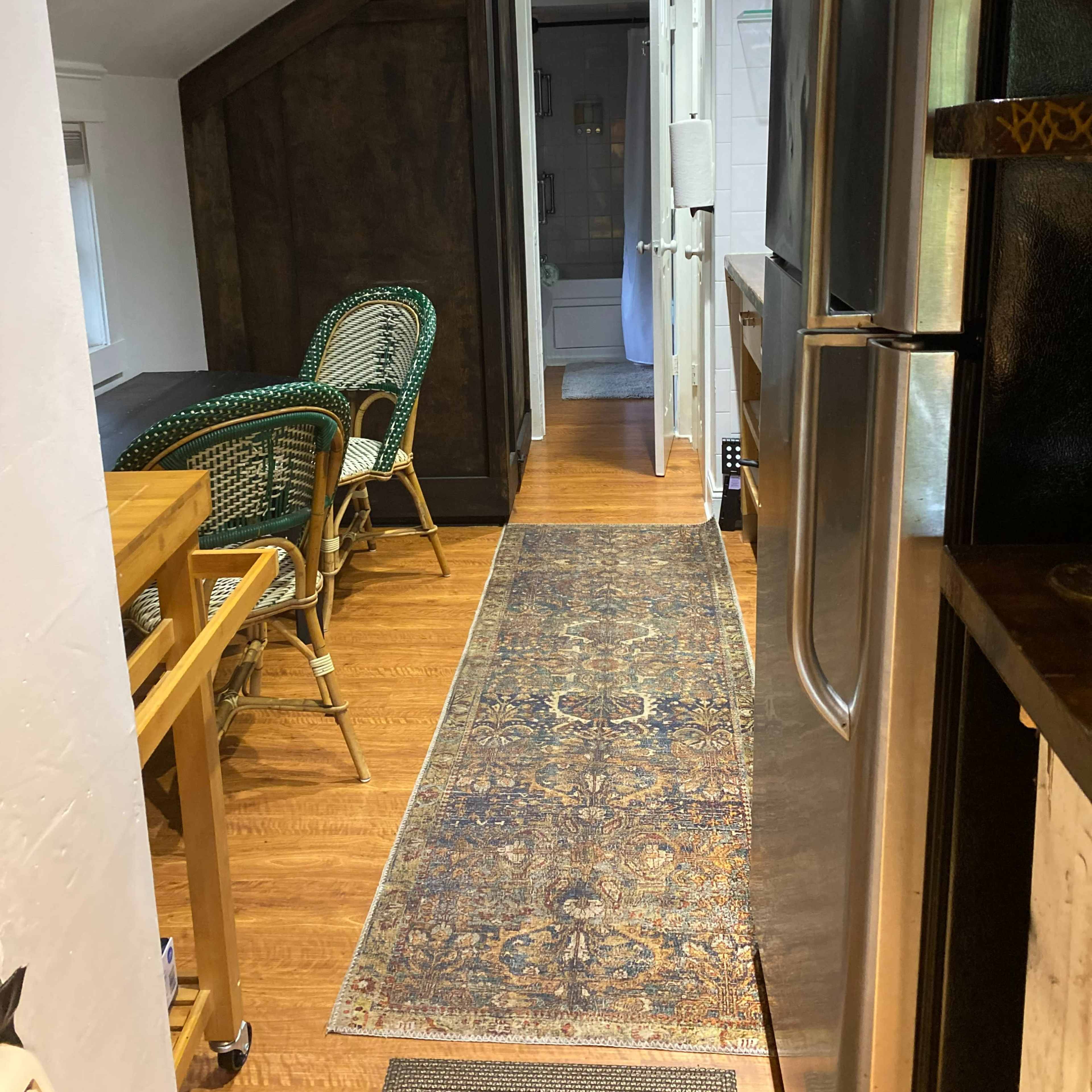 A narrow hallway in a home, featuring a kitchen with a stainless steel refrigerator, a dining table and chairs, and a doorway leading to a bathroom.