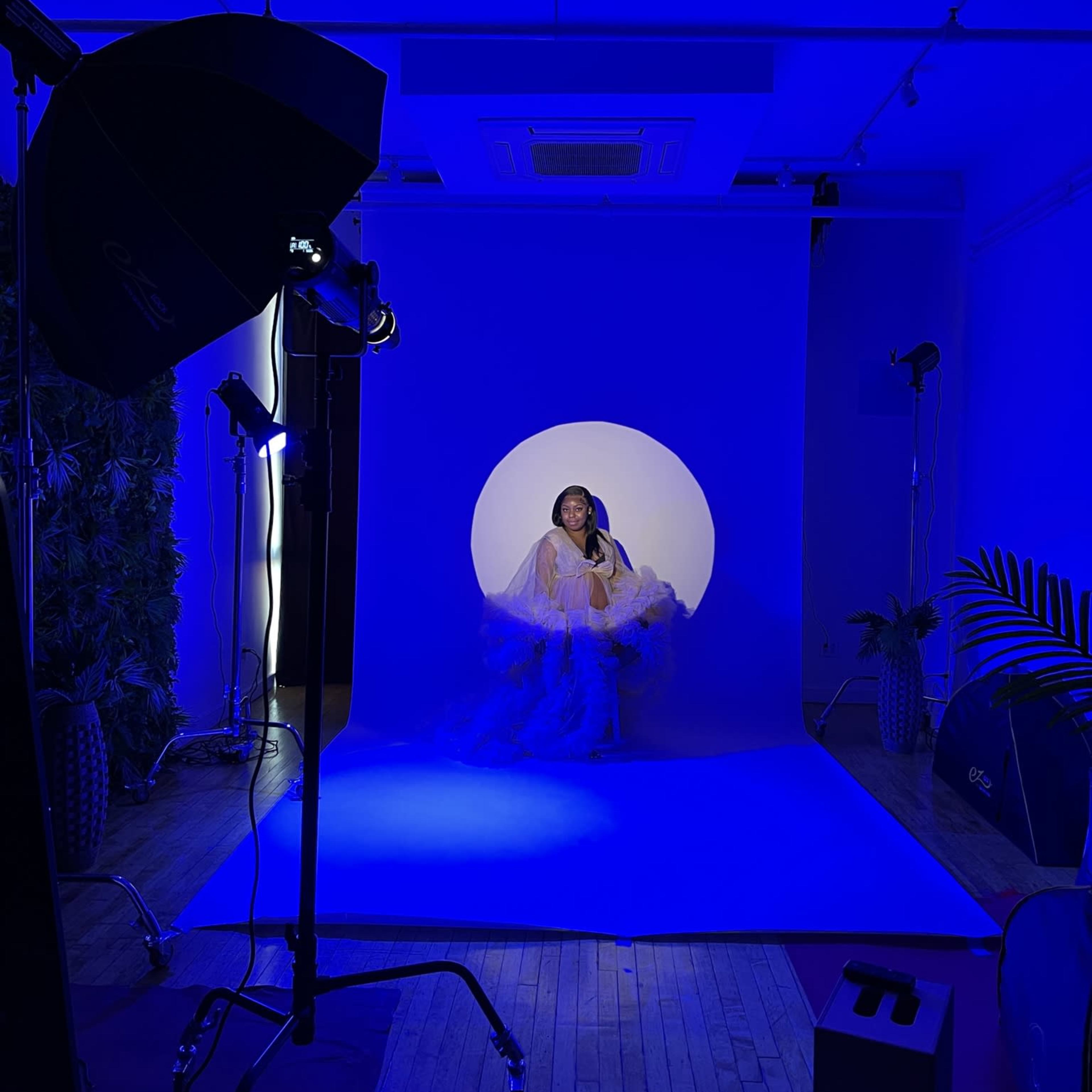 A woman sits on a blue backdrop with a circular cutout, surrounded by studio lighting and props.