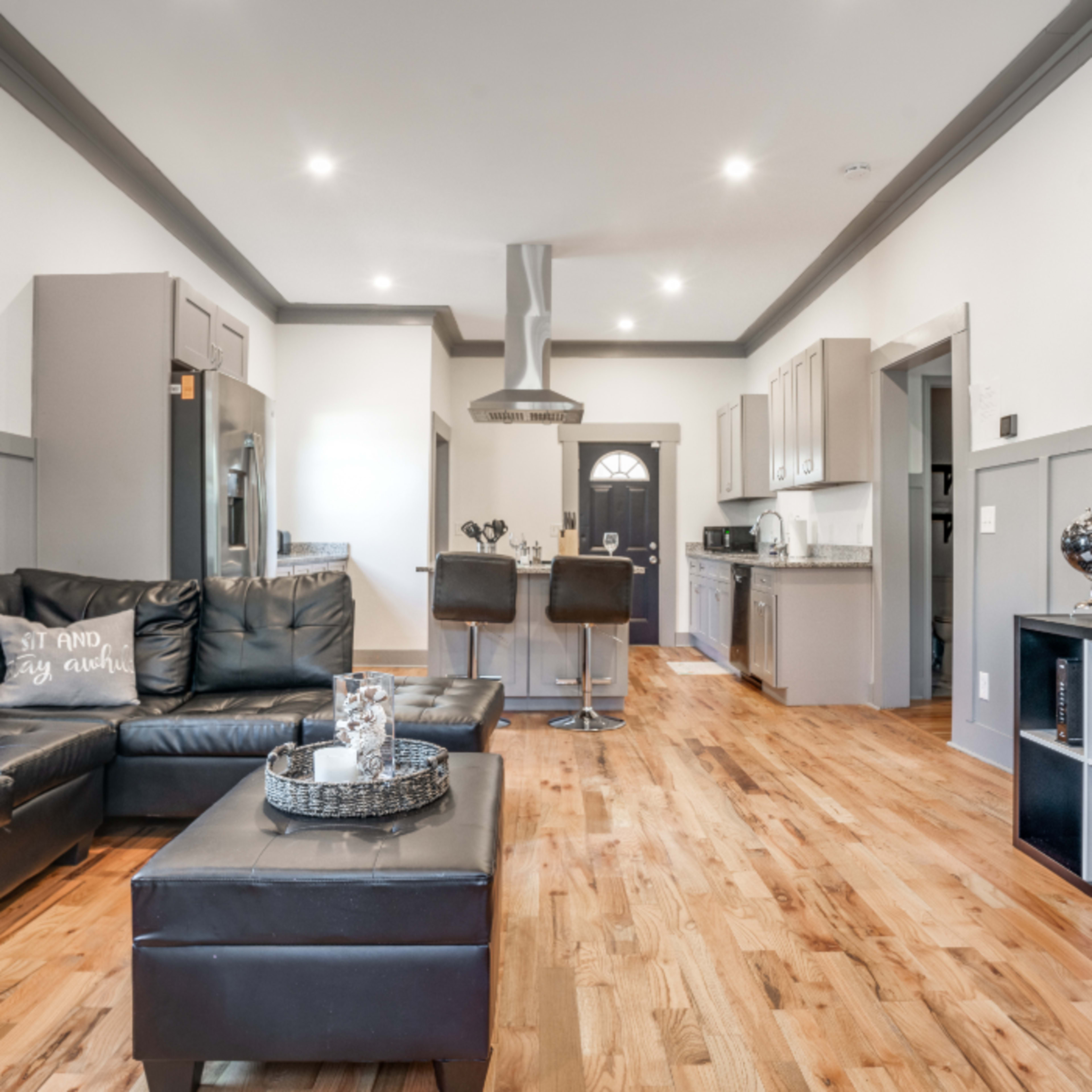 A modern living room with a black leather sectional sofa, a coffee table, and a kitchen area in the background featuring gray cabinetry and stainless steel appliances.