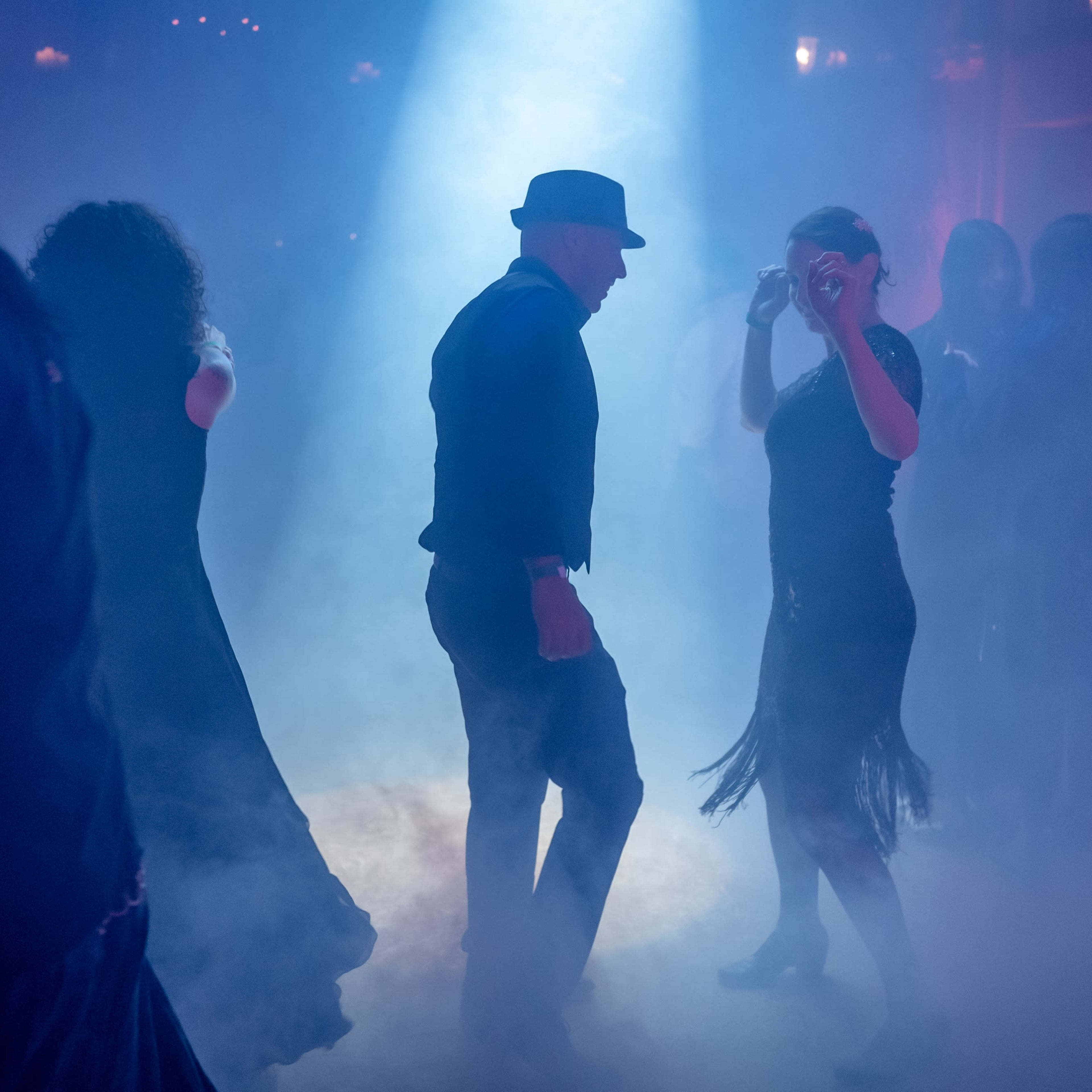 A group of dancers moves in a dimly lit space filled with smoke, highlighted by a beam of light.