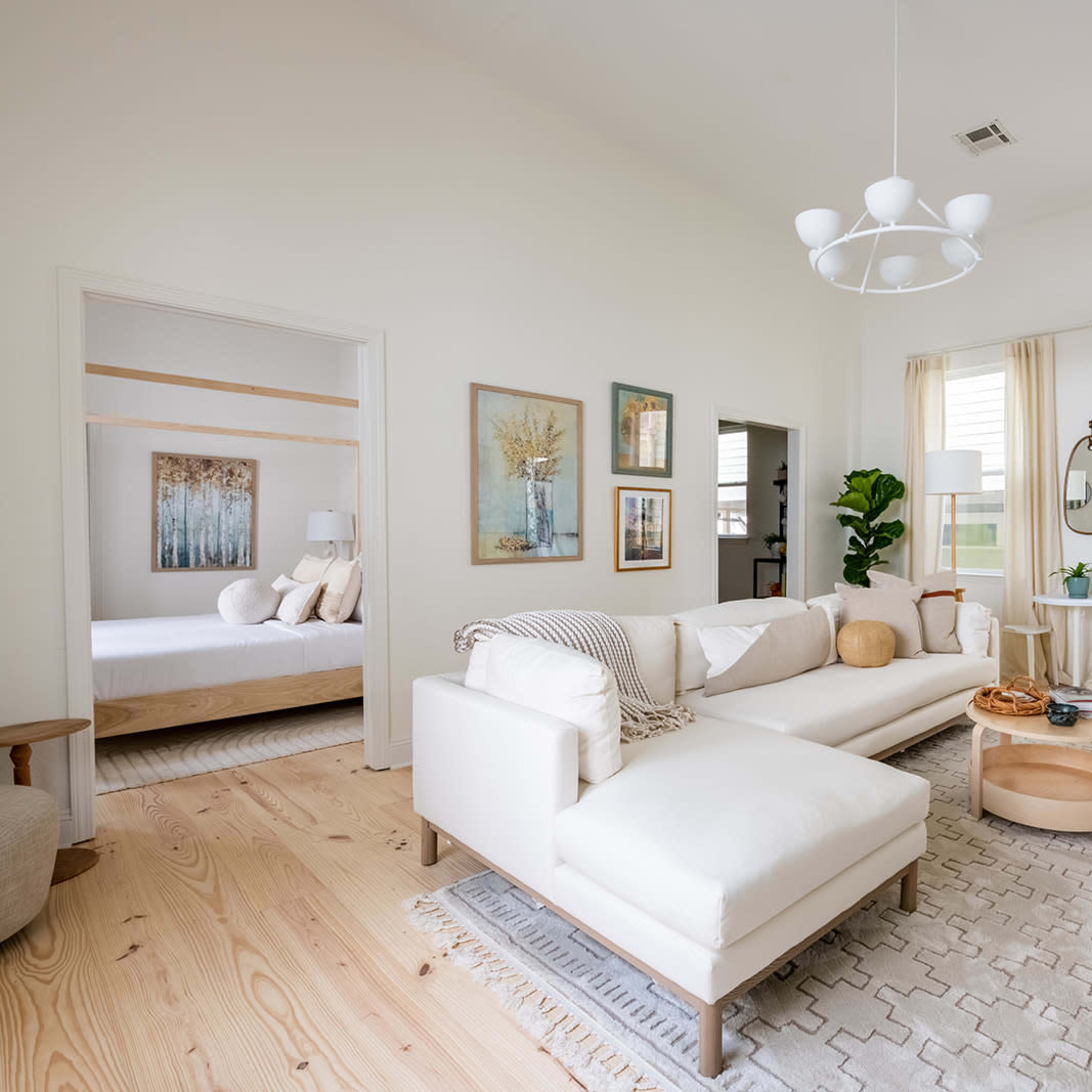 A spacious living room with a light color palette, featuring a white sectional sofa, a round coffee table, and large windows with sheer curtains.