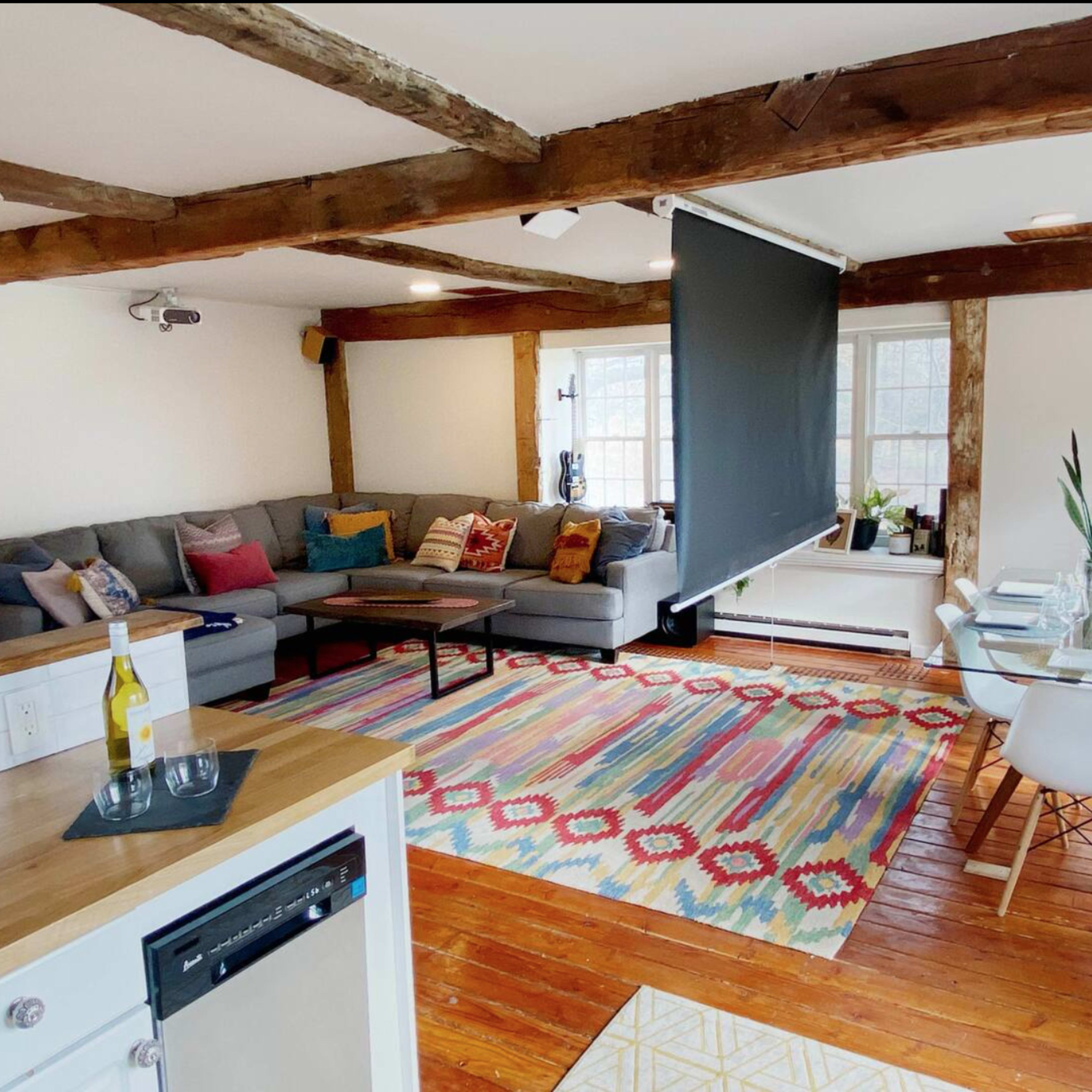 A spacious living area with a gray sectional sofa, a dining table for six, a colorful area rug, and wooden beams on the ceiling.