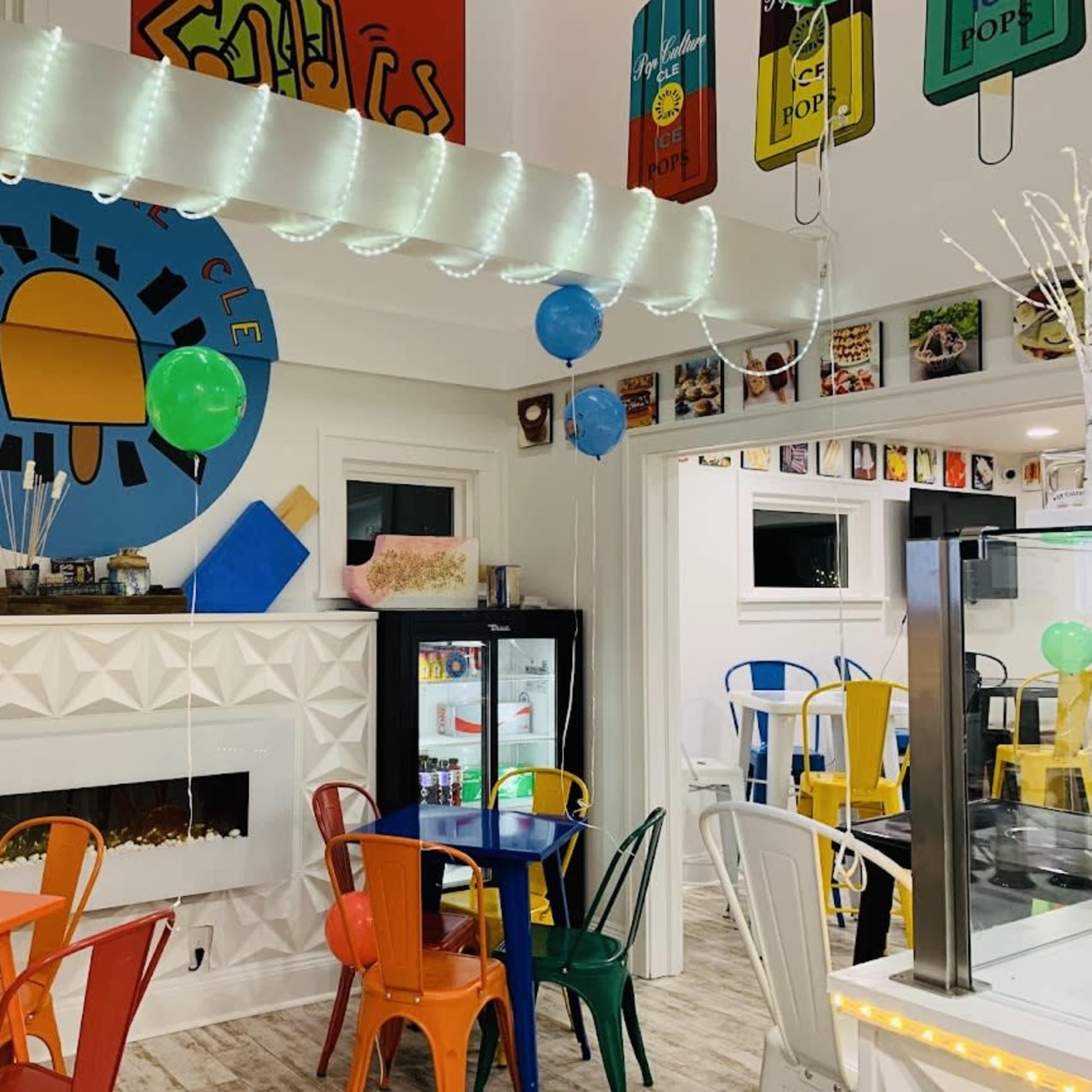 A colorful cafe interior with a decorative wall mural, assorted chairs around tables, and shelves displaying various items.