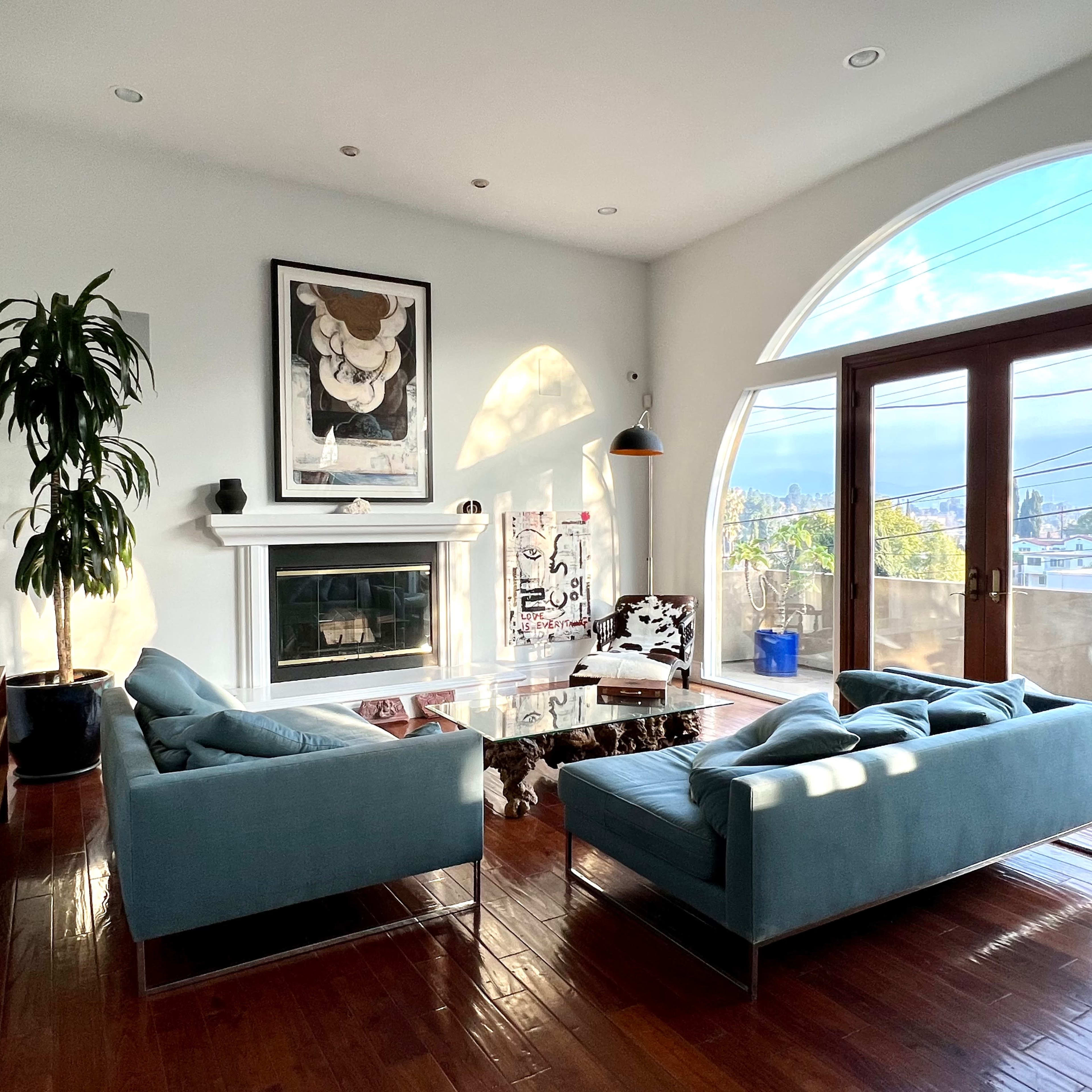 The living room features two blue sofas facing a glass coffee table in front of a white fireplace, with large windows offering a view of the outside.