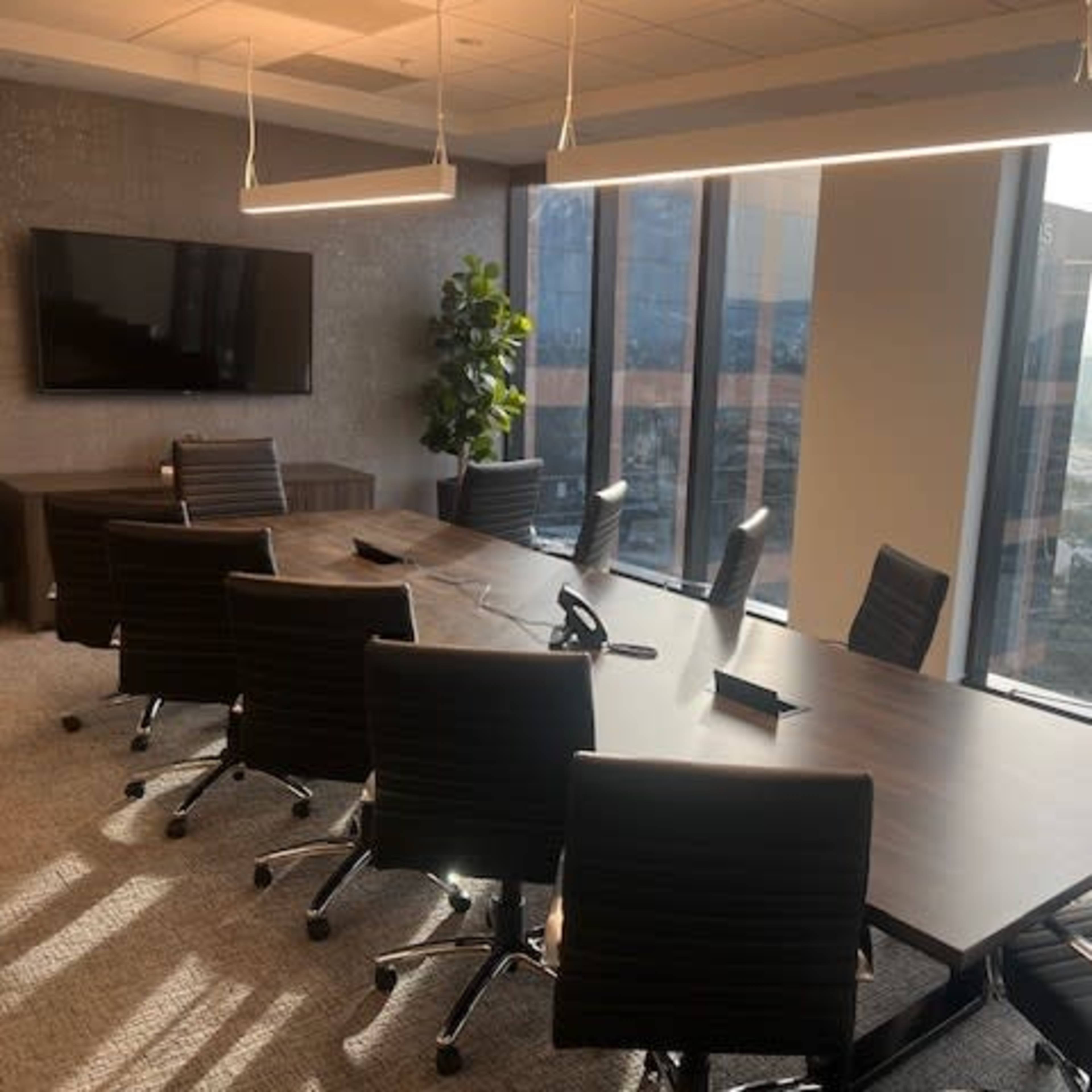 A modern conference room features a large wooden table surrounded by rolling chairs and large windows that offer a view of the city.