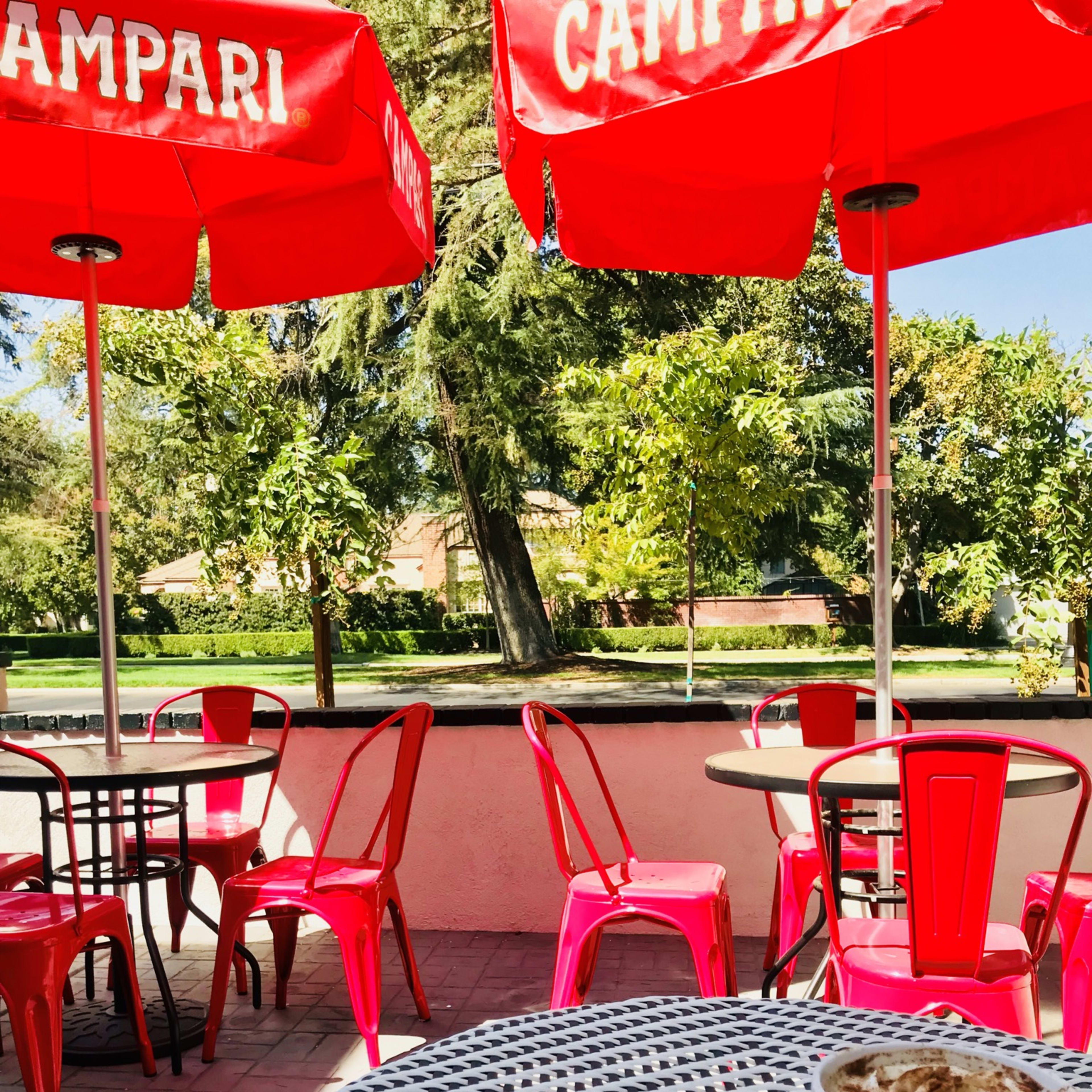 A table with a plate of cookies and a cup of coffee sits in a café with red umbrellas and outdoor seating in a park-like setting.