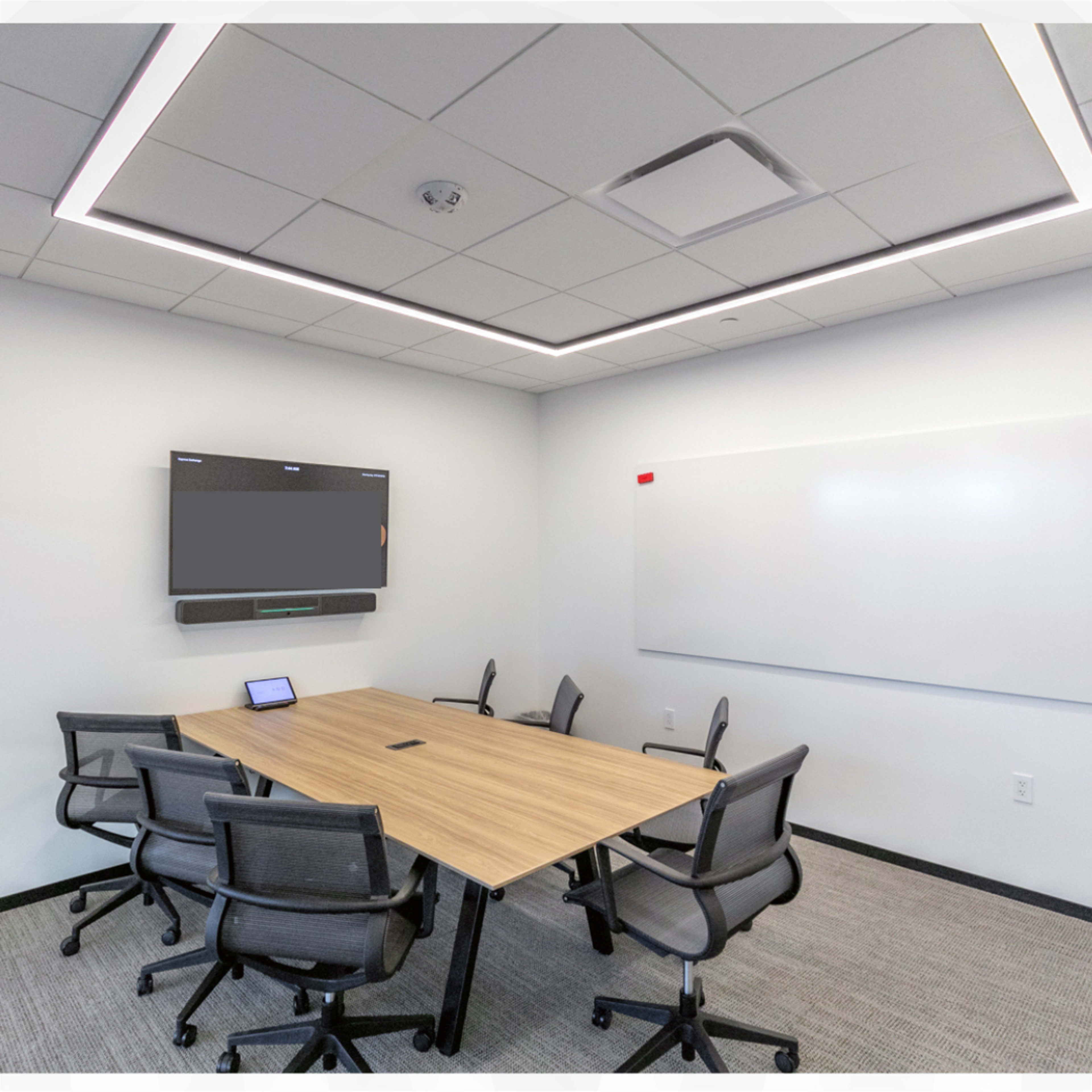 A meeting room featuring a rectangular wooden table surrounded by six office chairs, with a flat-screen TV on the wall and a whiteboard nearby.