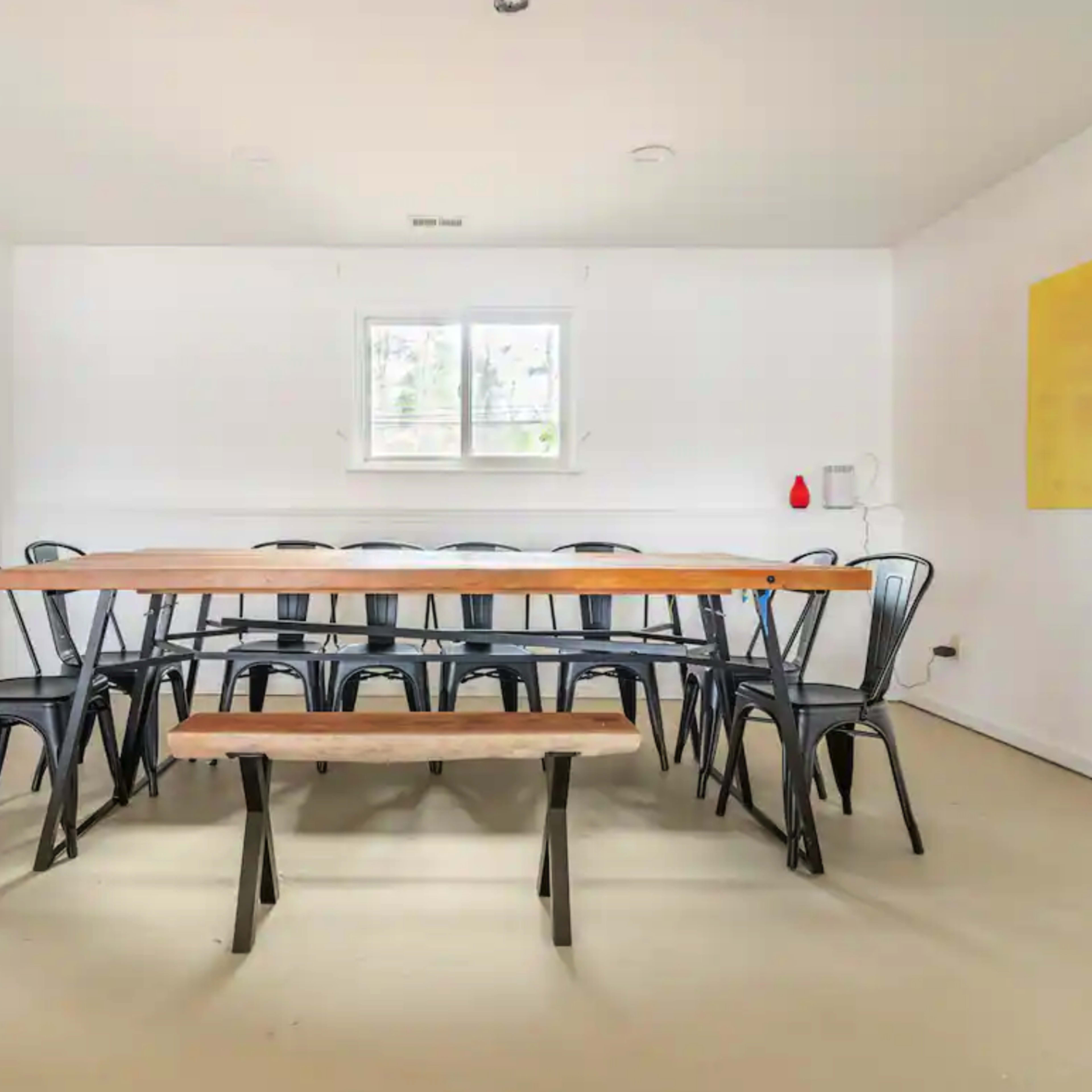 A spacious dining area featuring a long wooden table surrounded by black metal chairs, with a bench on one side and minimalist decor on the walls.