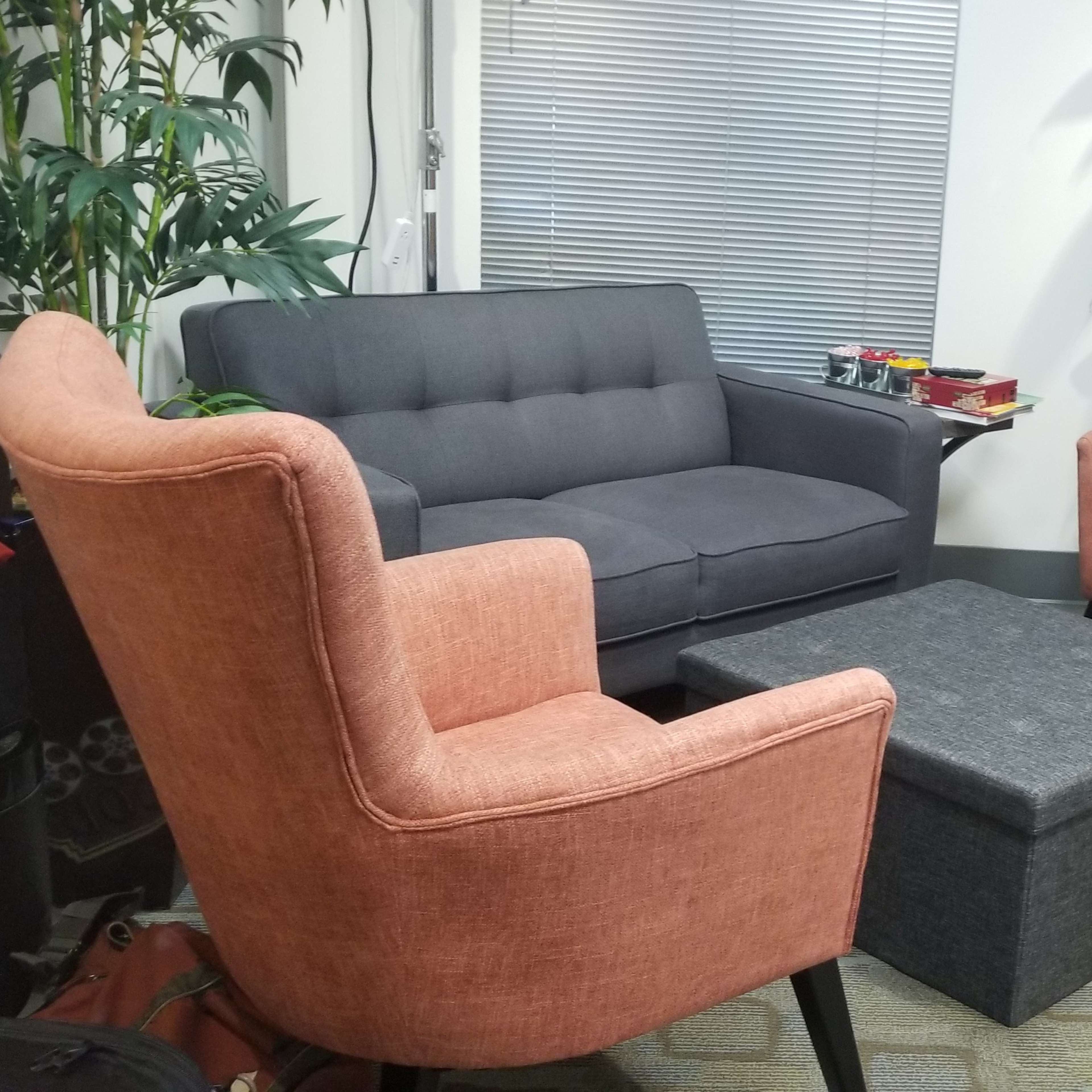 A small sitting area with a gray sofa and two pink armchairs surrounding a gray ottoman, with plants and a window in the background.