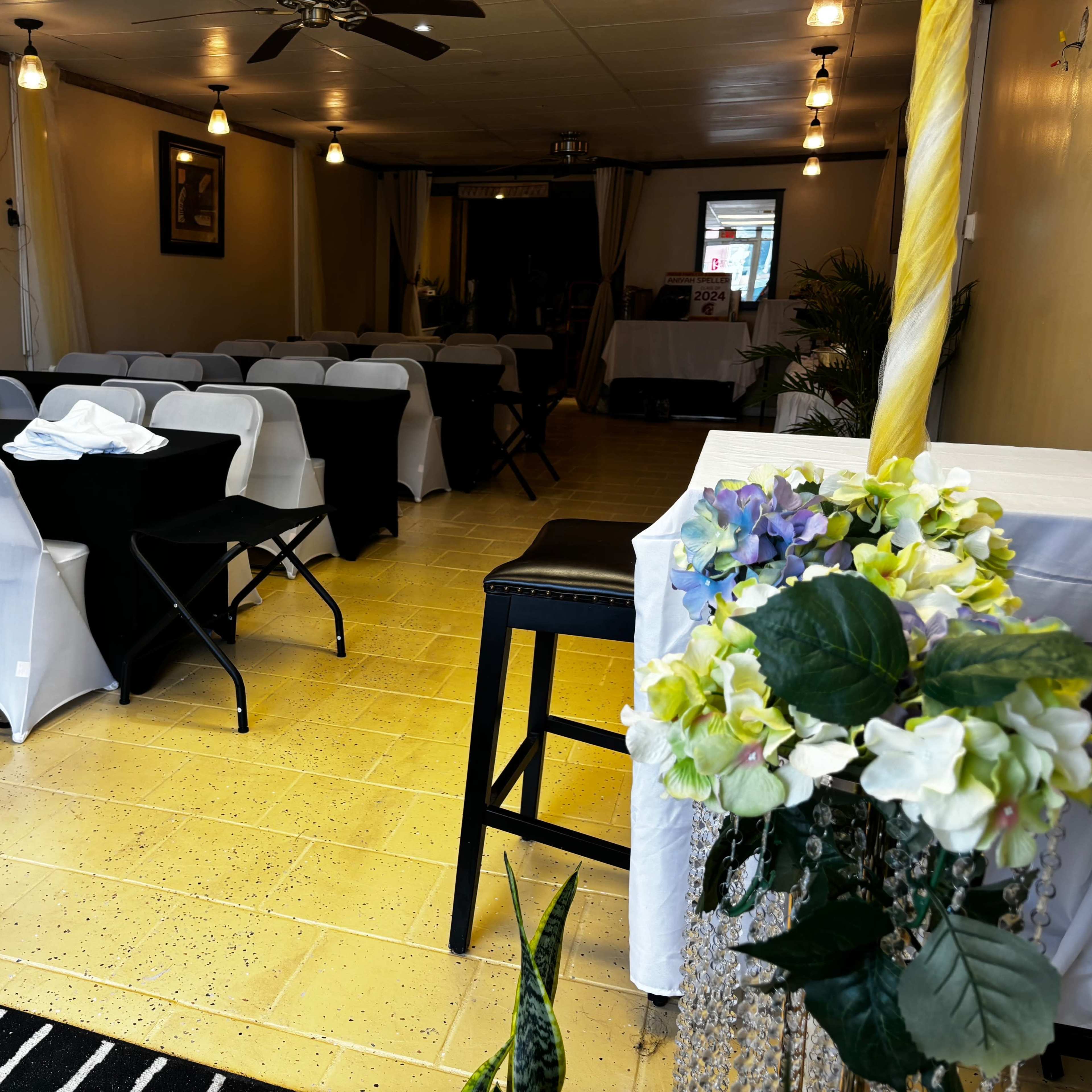 A decorated indoor event space with rows of tables covered in black and white cloths, and a floral arrangement on a nearby table.