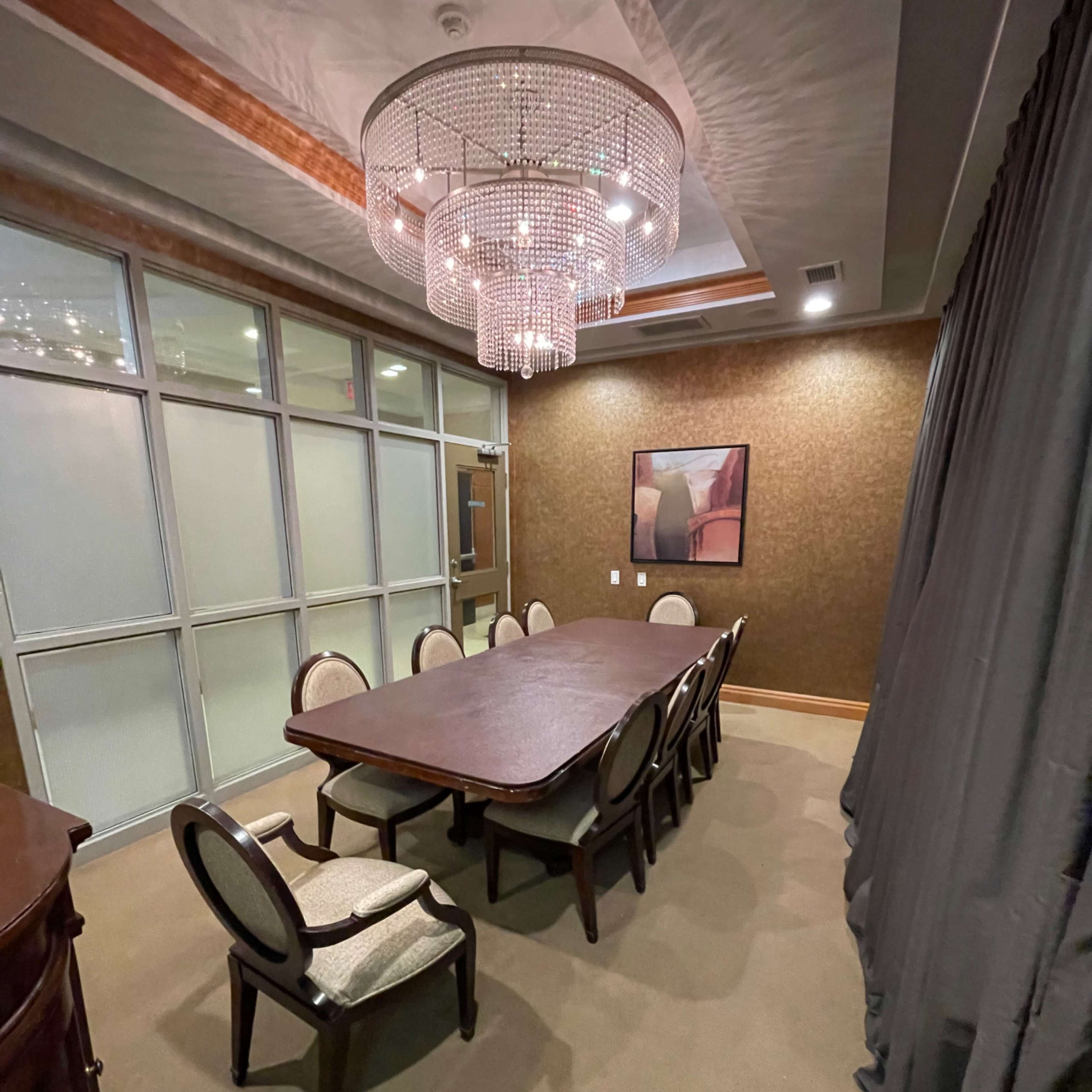 A conference room with a large wooden table surrounded by chairs, a chandelier overhead, and a wall covered in textured fabric.