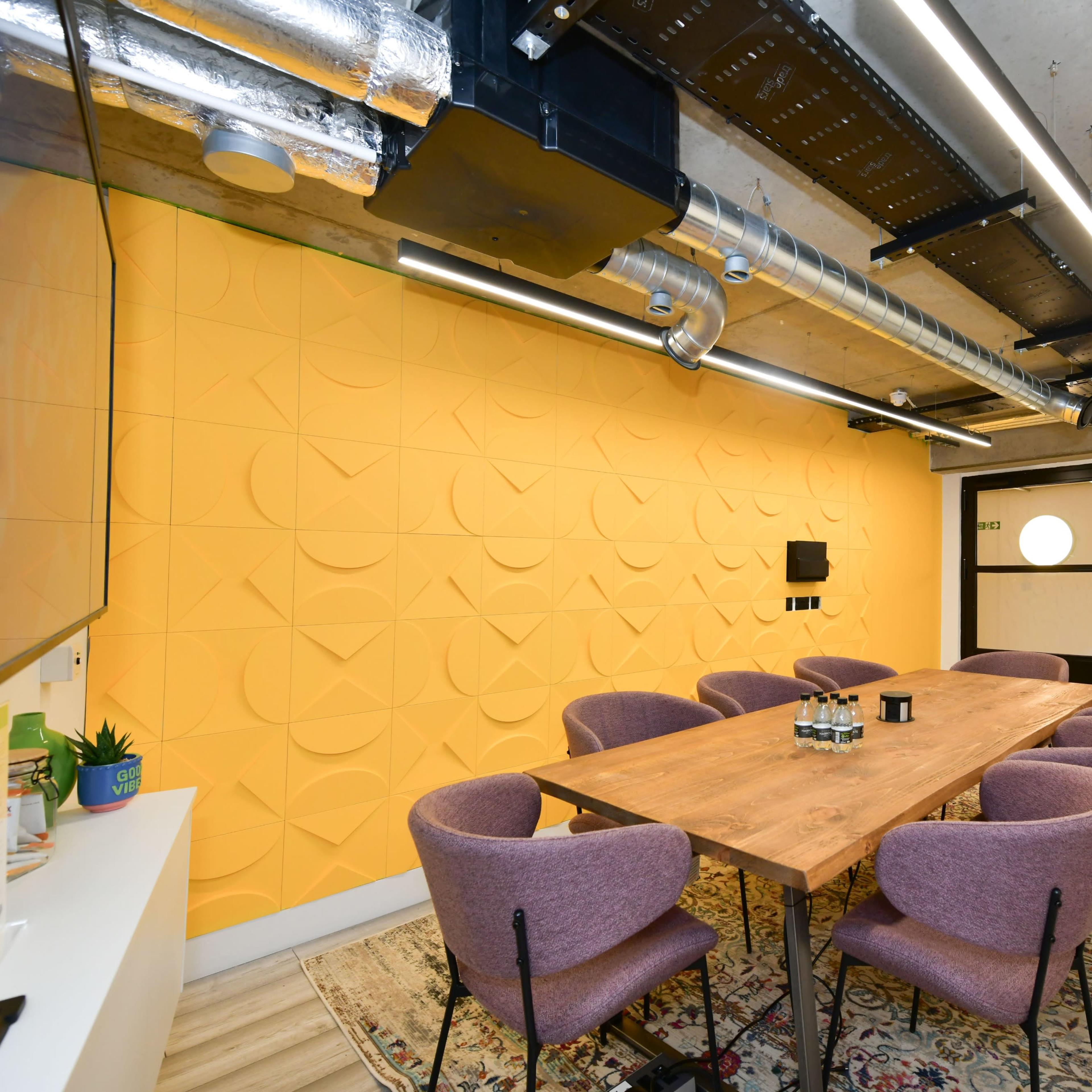 A modern meeting room with a long wooden table surrounded by purple upholstered chairs, complemented by a bright yellow textured wall and exposed ceiling elements.