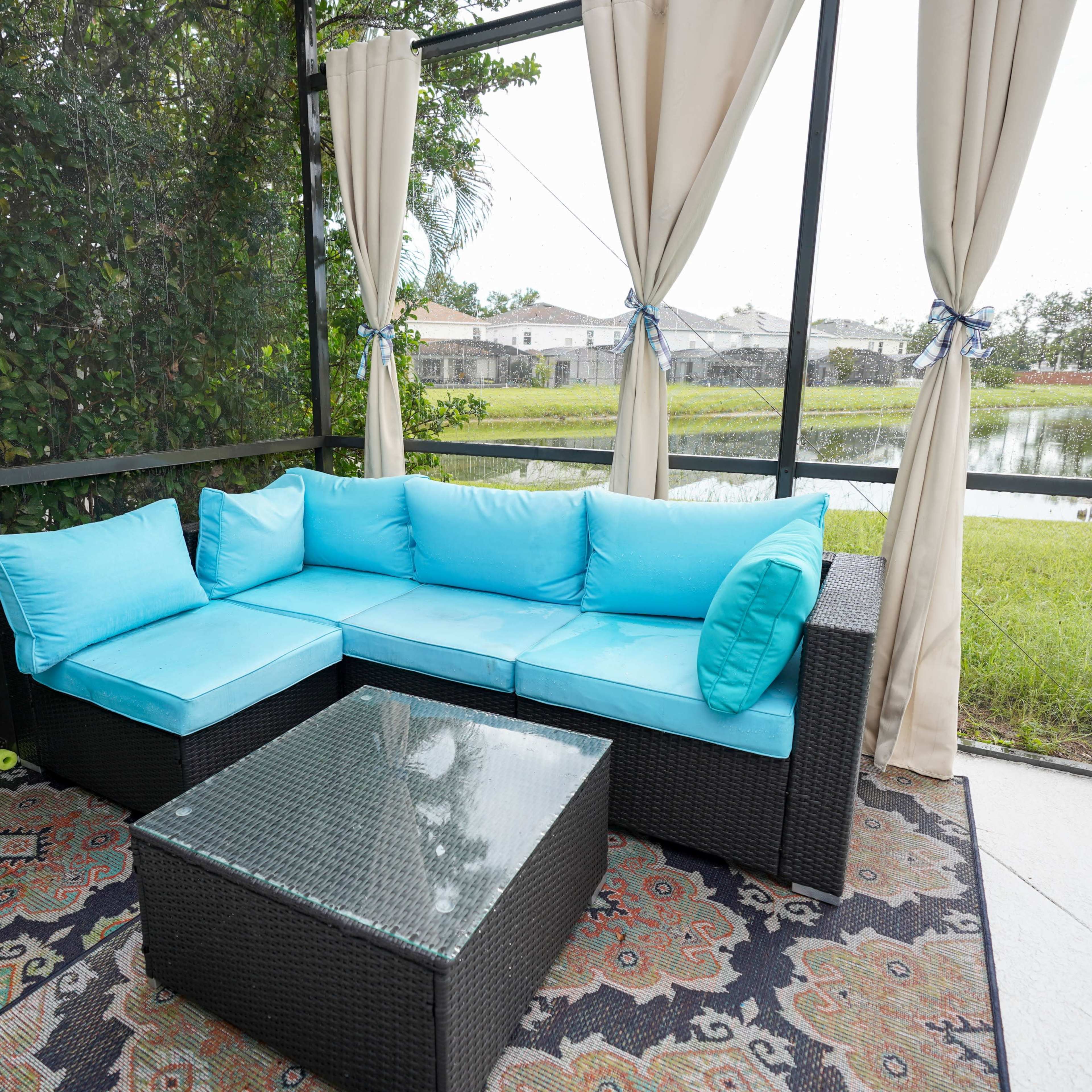 A patio seating area features a light blue sectional sofa and a glass-topped coffee table, surrounded by greenery and a view of a pond.