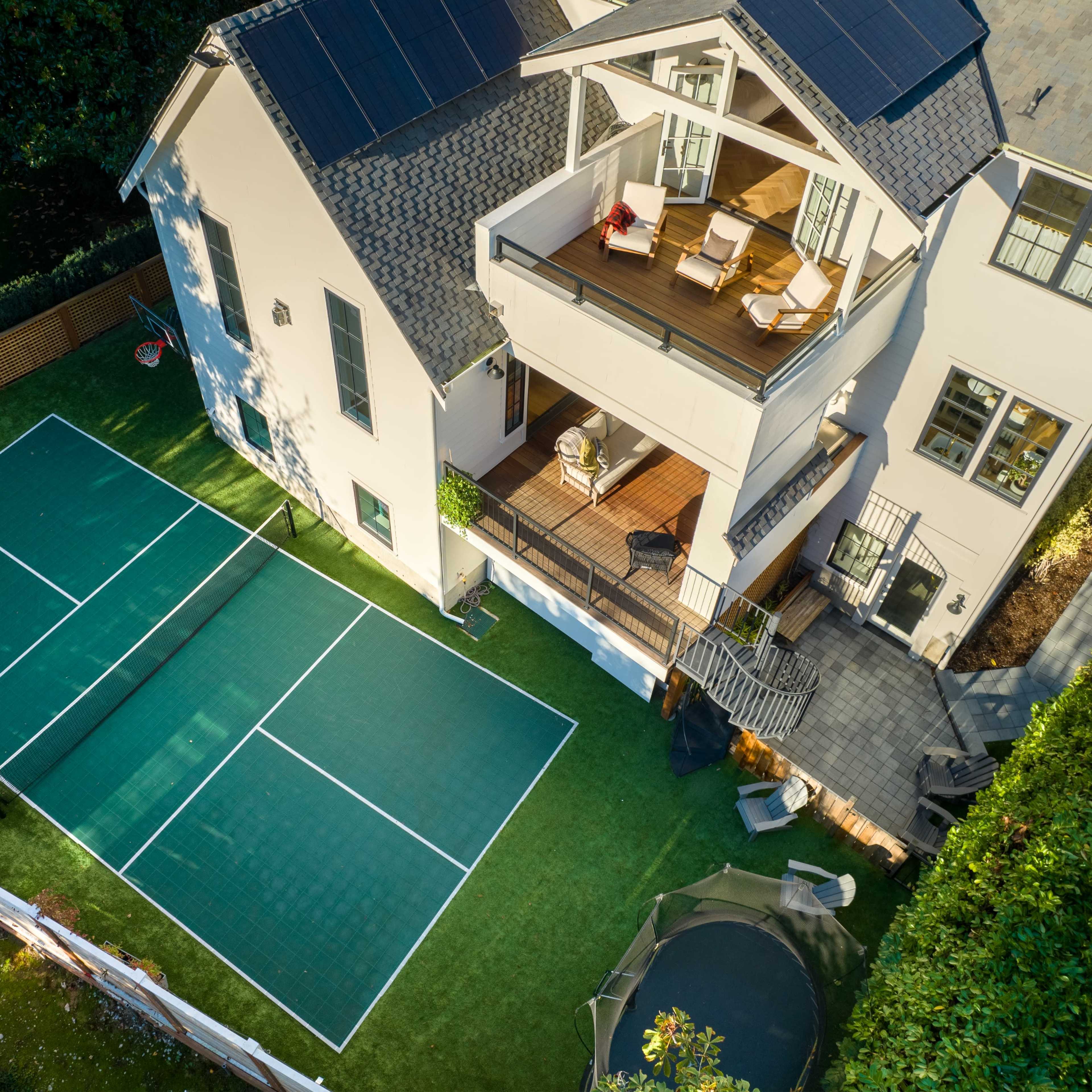 A two-story house with solar panels on the roof, a balcony with seating, and a green tennis court in the backyard.