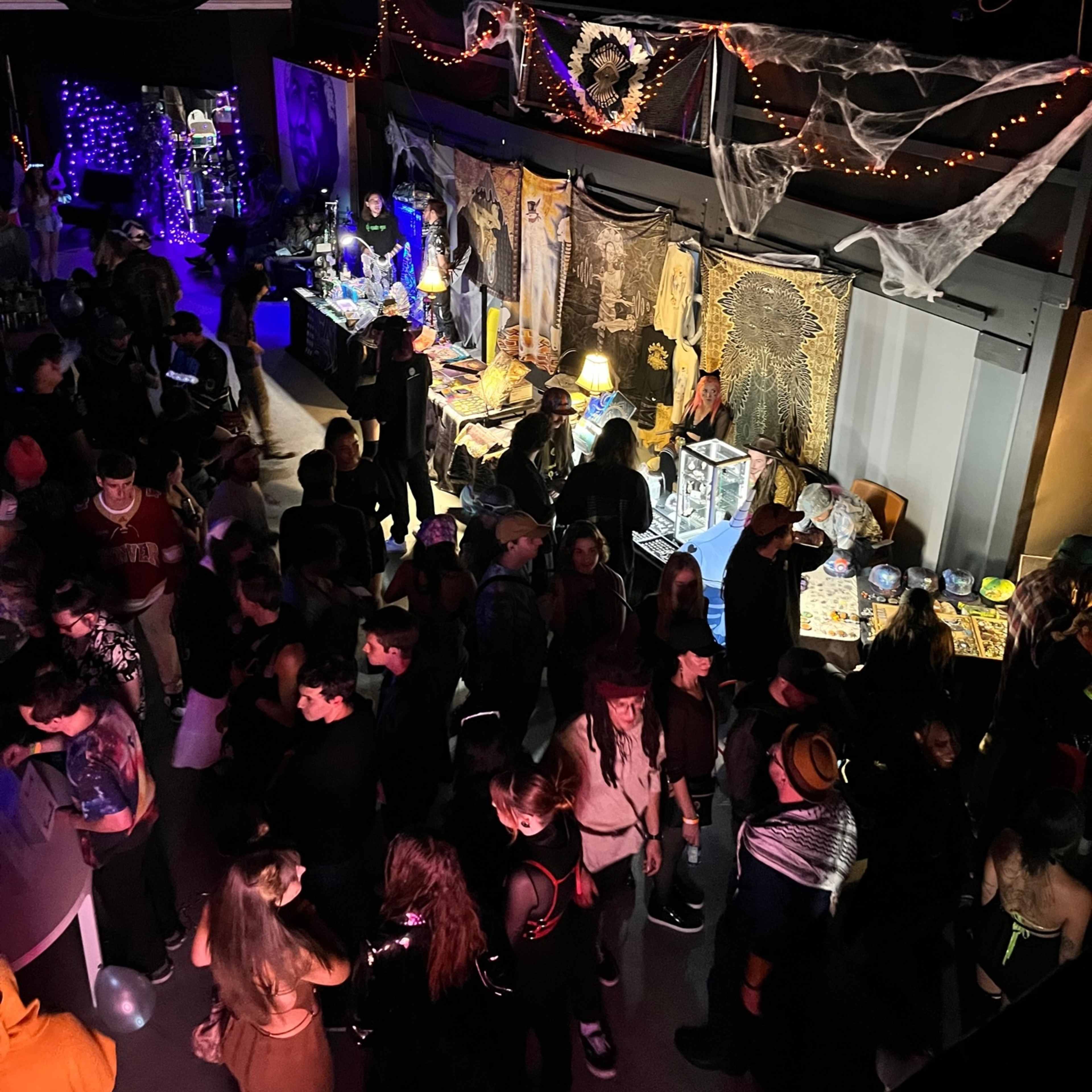 A crowd of people gathers in a dimly lit party venue decorated with spider webs and various props.