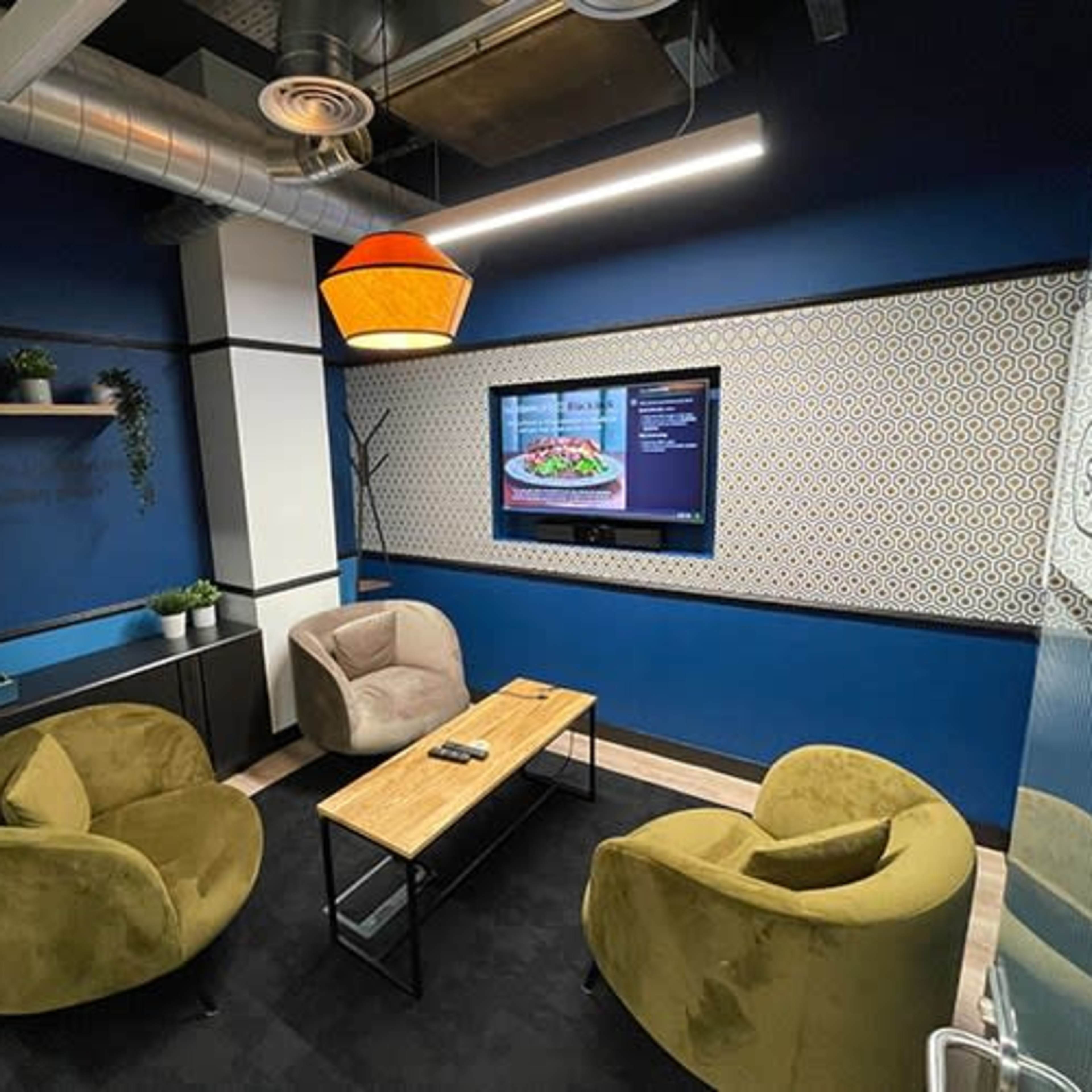 A cozy meeting room features two armchairs and a coffee table, with a television displaying a menu on a blue wall.