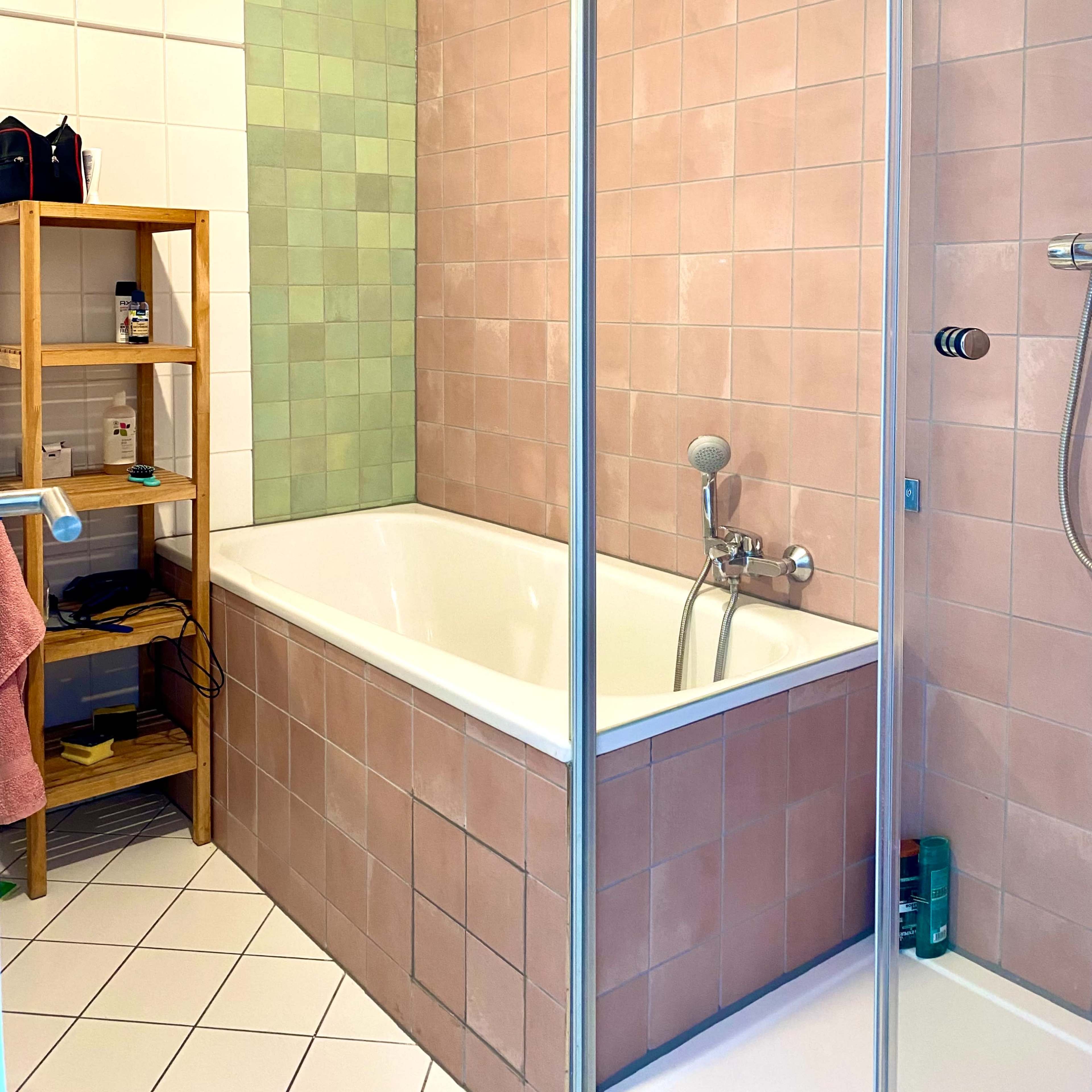 A bathroom with a bathtub and shower, featuring tiled walls in pink and green, along with a wooden shelf holding various toiletries.