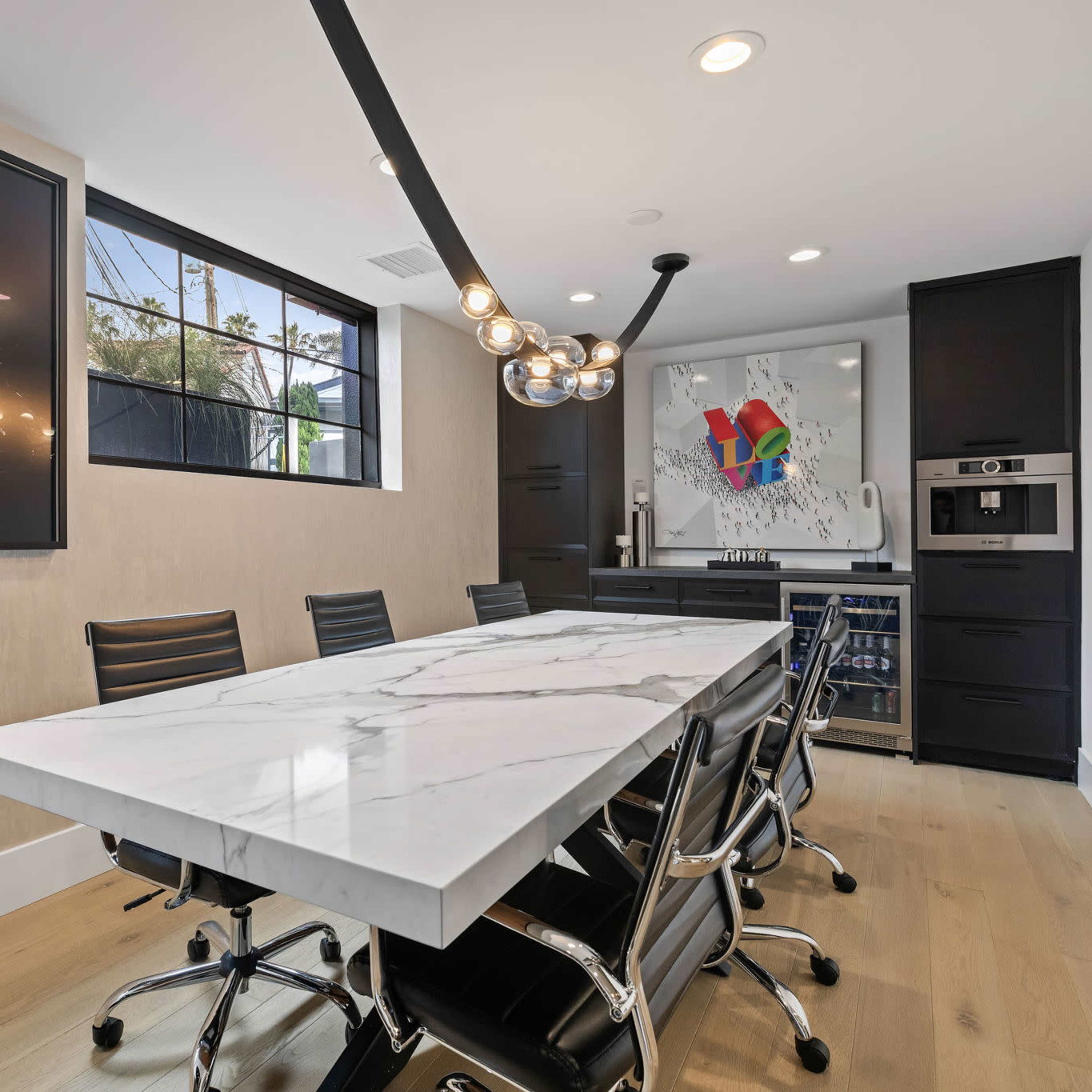 A modern dining area features a long marble table surrounded by black chairs, with large windows and artwork on the walls.