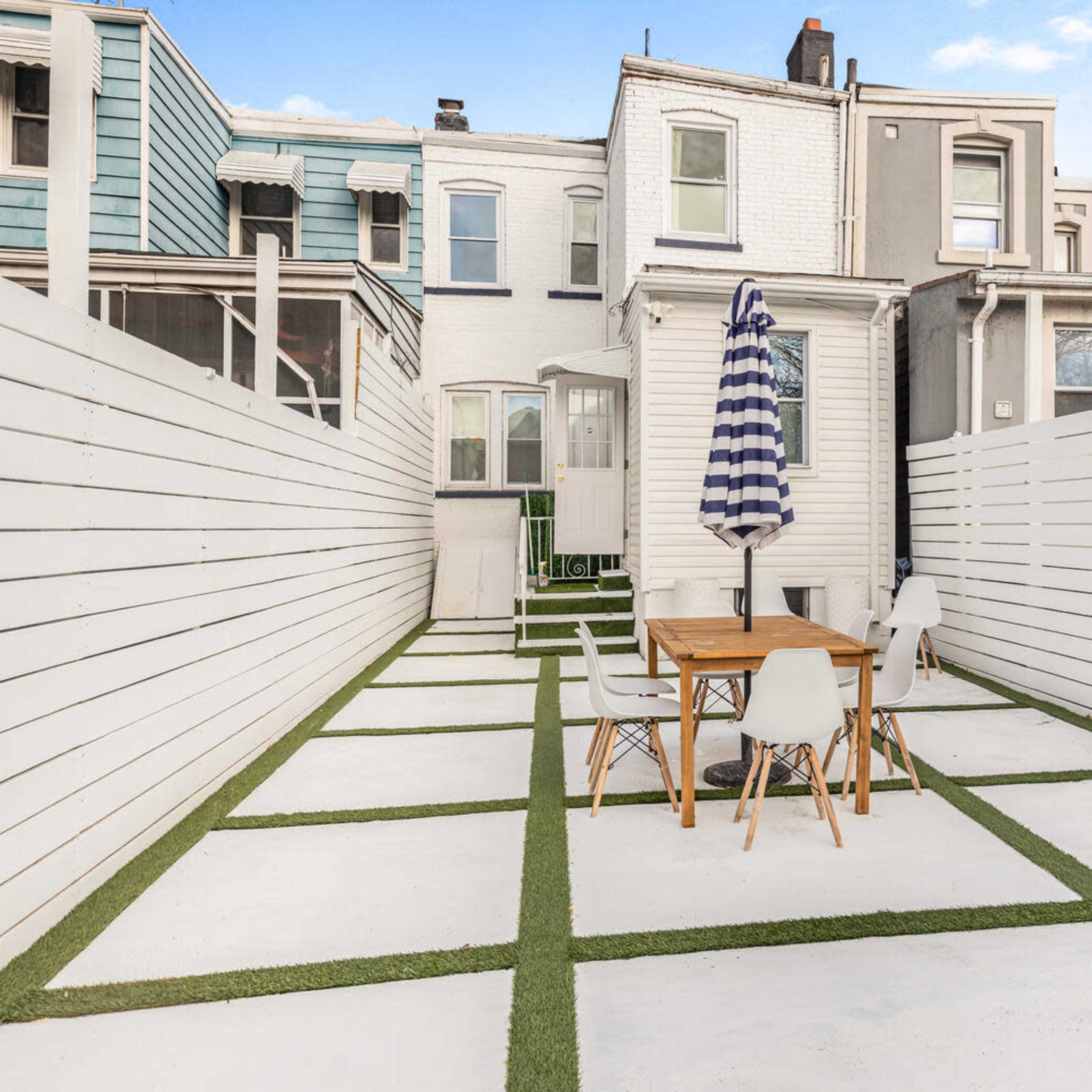A small outdoor patio area with a table and chairs, flanked by white fences and covered with artificial grass accents.