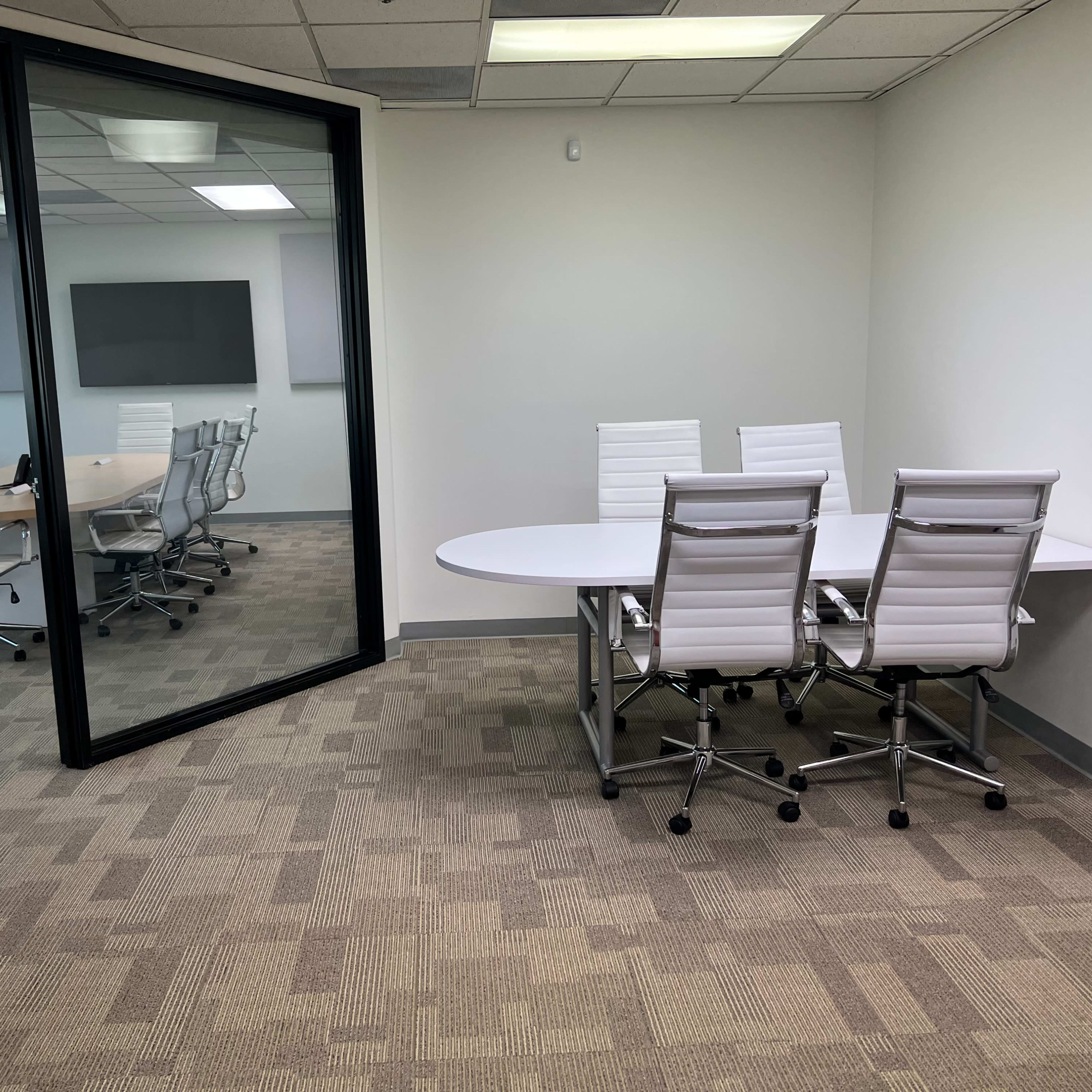 A modern office space with a glass-walled conference room and a separate table with white chairs in an adjacent area.