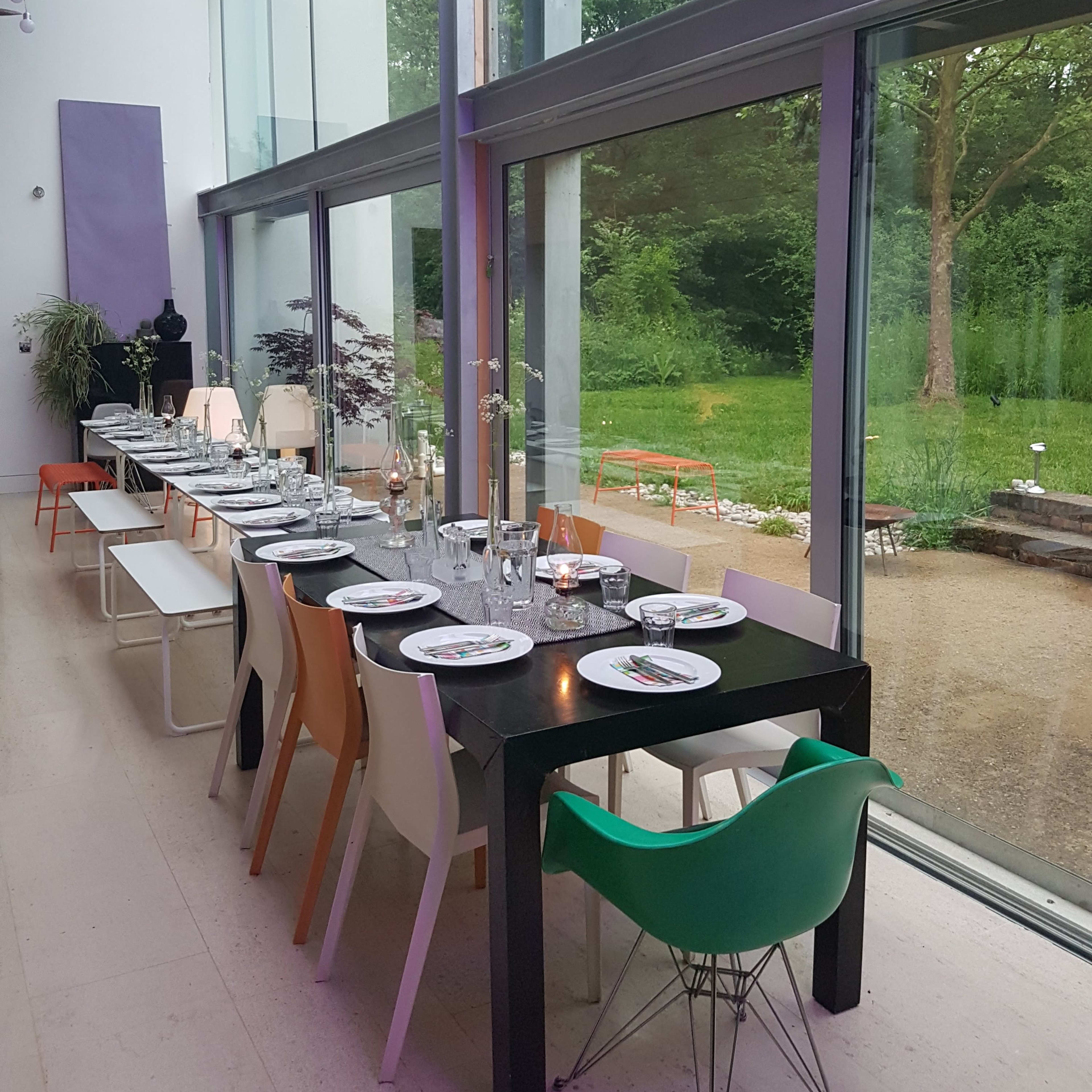 A long dining table is set with plates and glassware in a modern room featuring large windows that overlook a green garden.