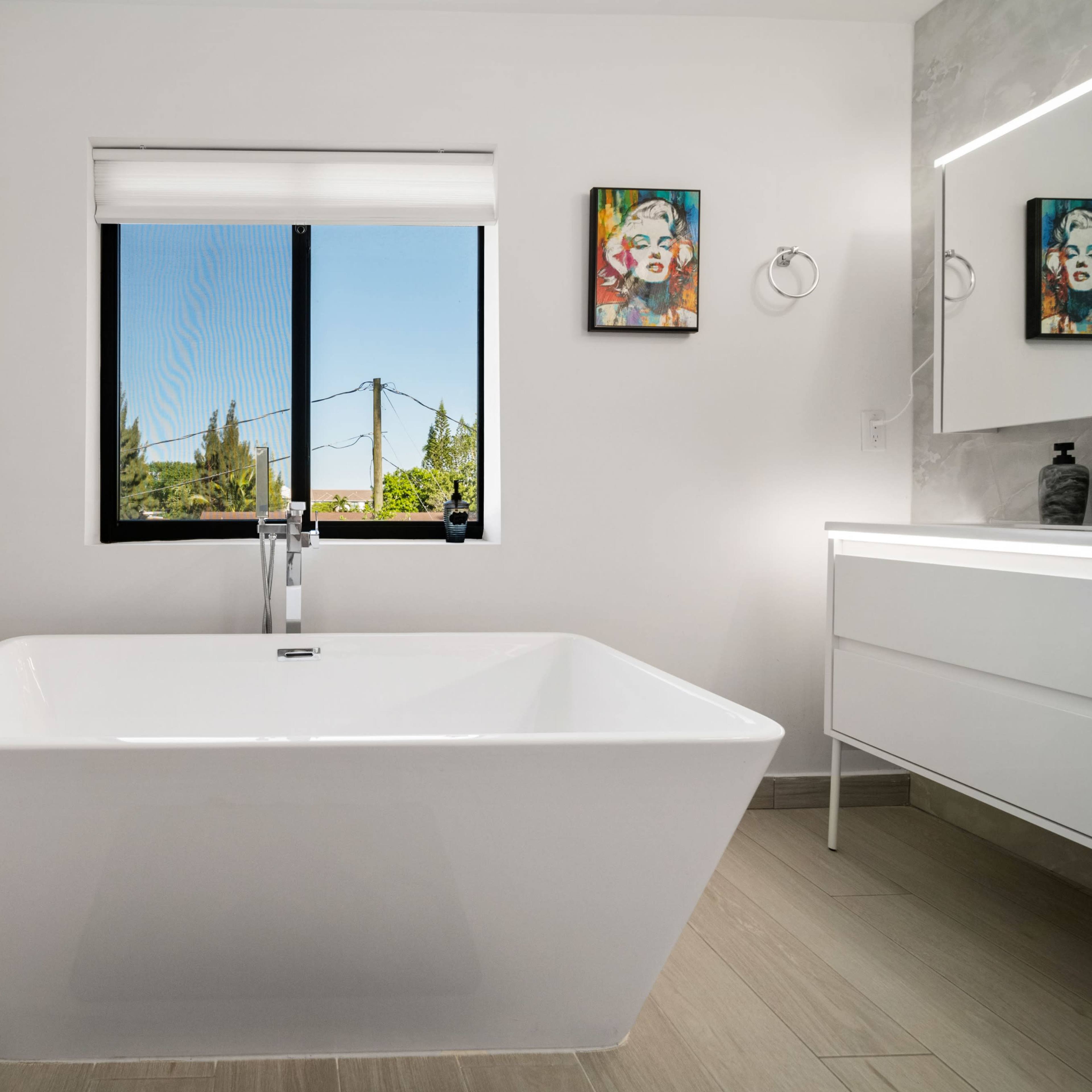 A modern bathroom features a freestanding white bathtub beneath a window with a view, alongside a wall-mounted mirror and artwork.