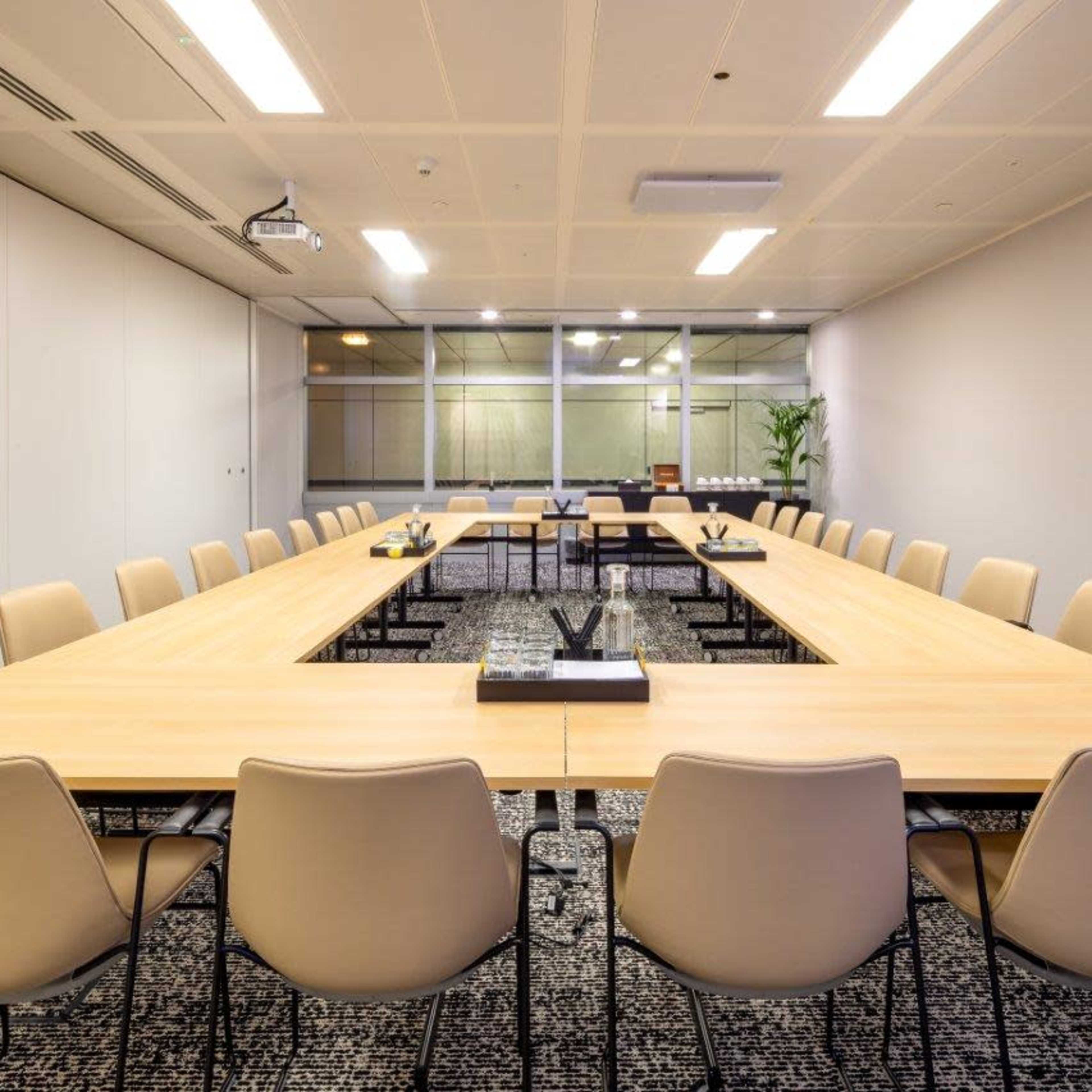 A large, rectangular conference room features a long wooden table surrounded by beige chairs, with a projector and glass windows in the background.