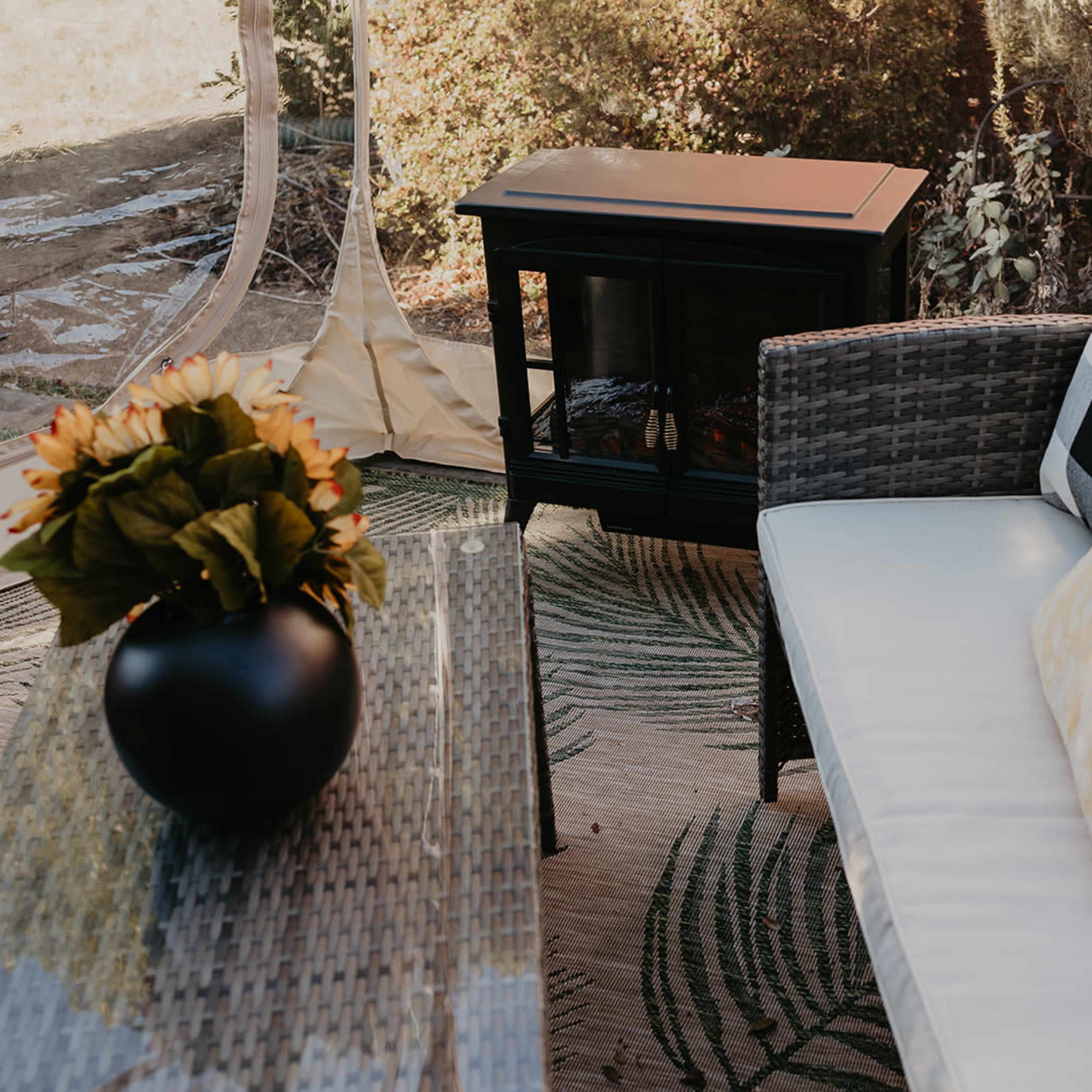 A cozy outdoor seating area features a wicker sofa with cushions, a decorative table, and a portable heater, all set within a transparent tent.
