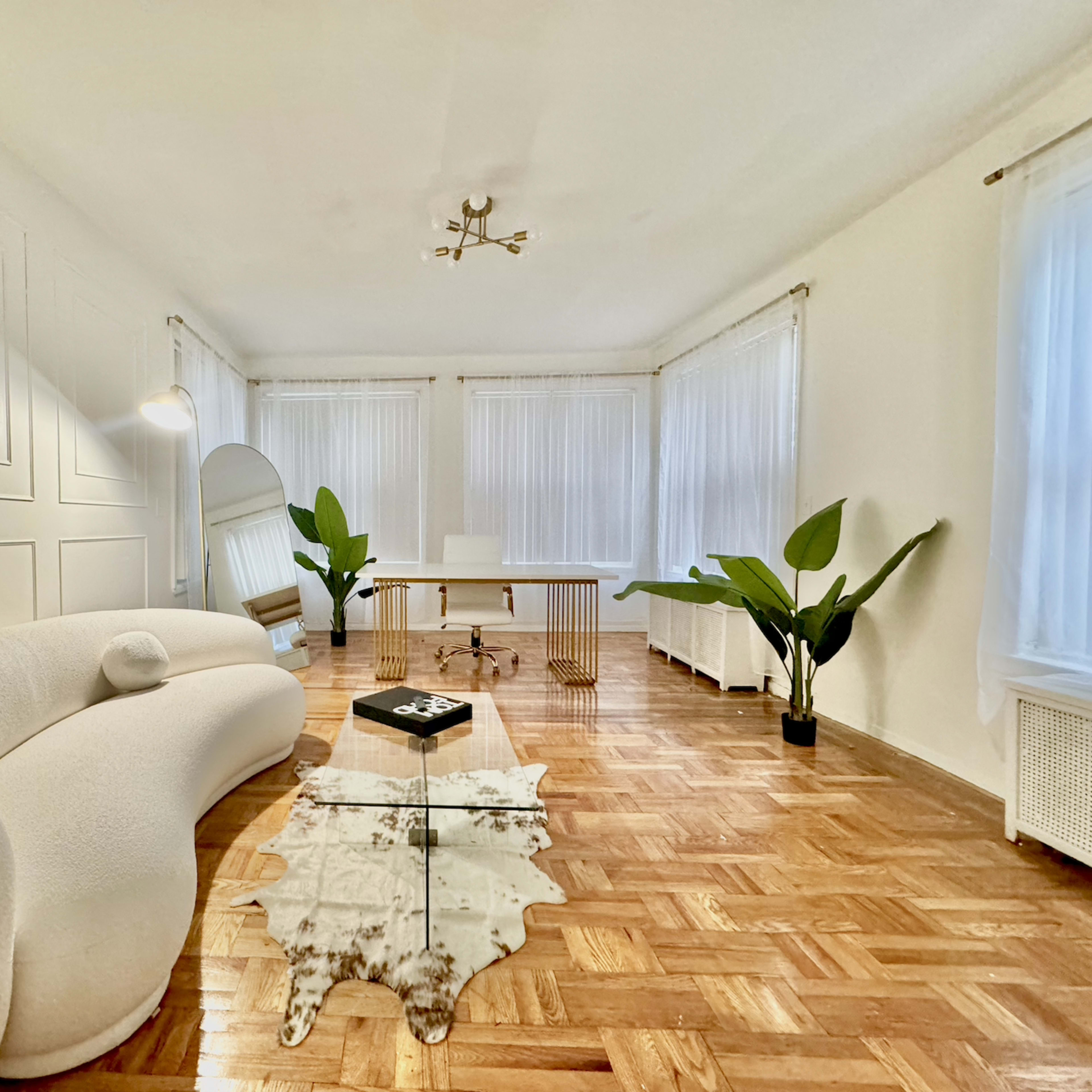 A spacious, well-lit room features a curved white sofa, a wooden desk, and decorative plants, all complemented by light-colored walls and large windows.