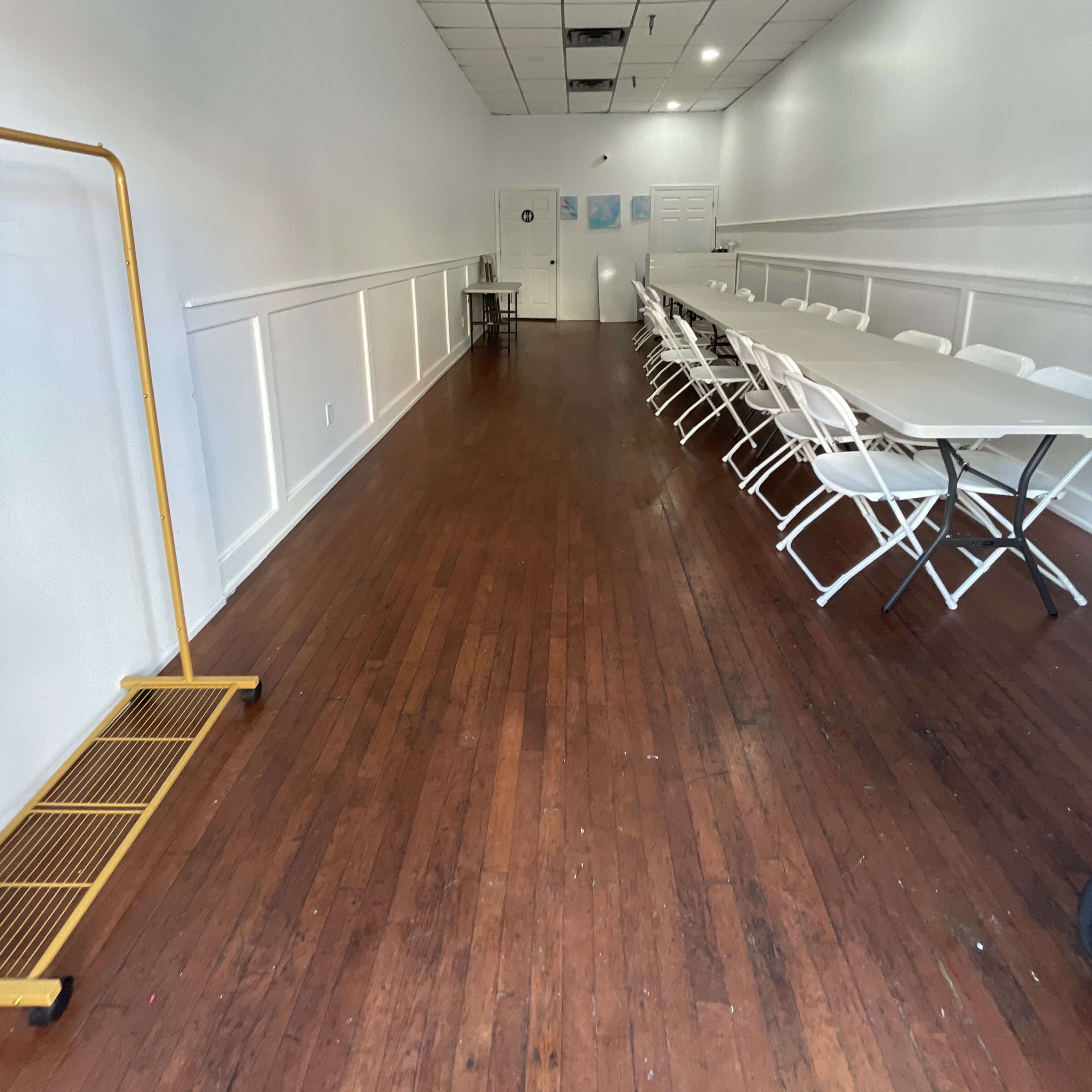 A long, narrow room with a wooden floor, features a row of white folding chairs and a long table, and has a rack along one side.